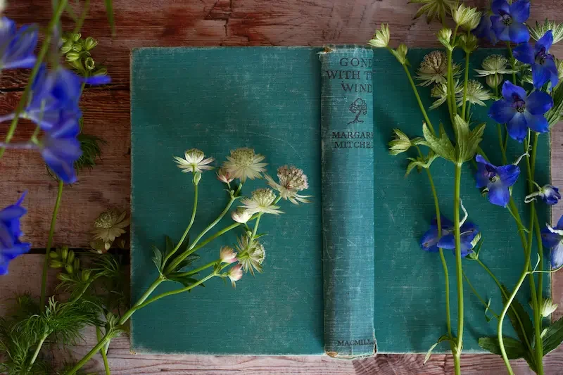 An open vintage teal book titled 'Gone with the Wind' by Margaret Mitchell, surrounded by white and blue flowers on a wooden surface.