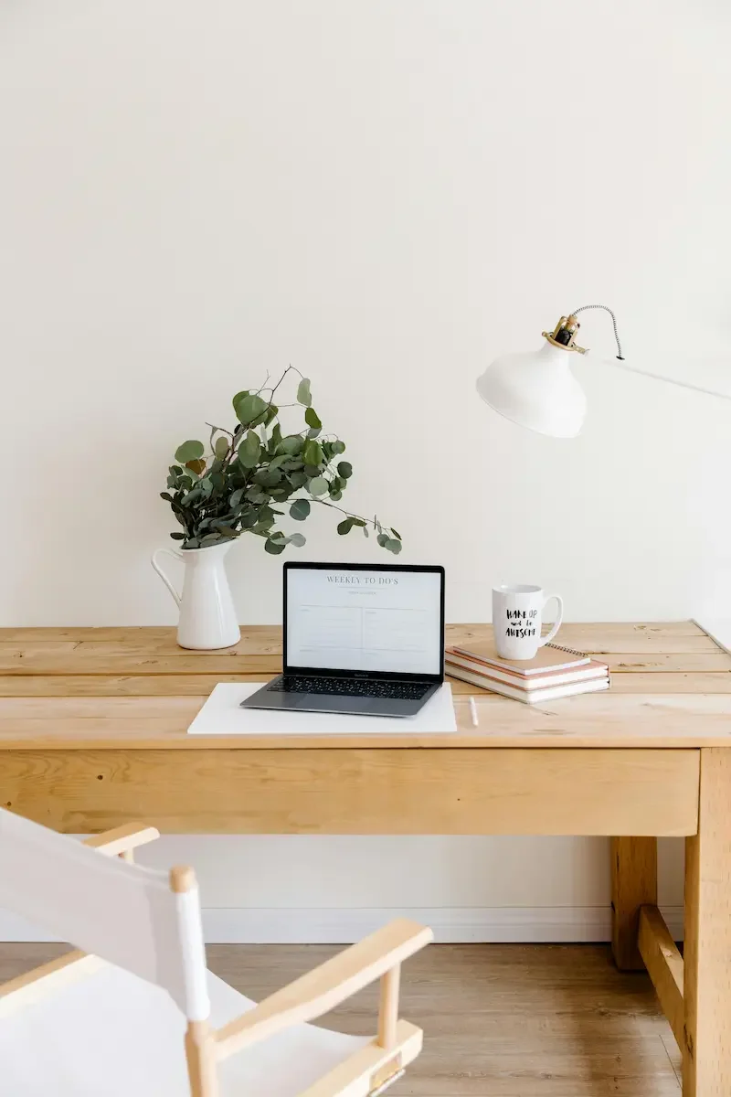Home workspace with a wooden desk holding a laptop, a white mug, a stack of books, and a potted plant, with a white adjustable desk lamp on the wall above.