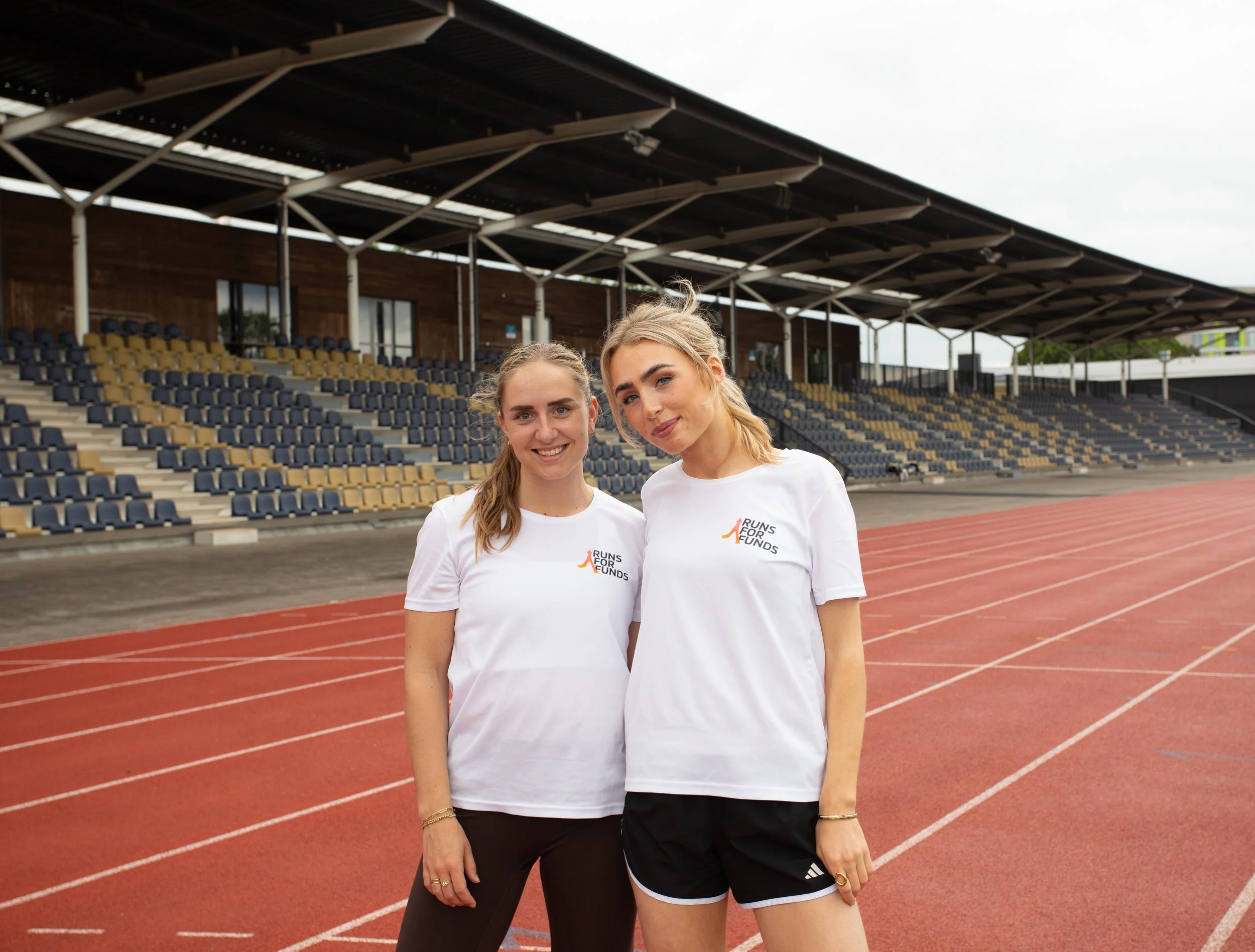 Twee jonge vrouwen staan op een atletiekbaan, langs een vol stadion met lege tribunes, en dragen witte T-shirts met 'Runs for Funds' logo.