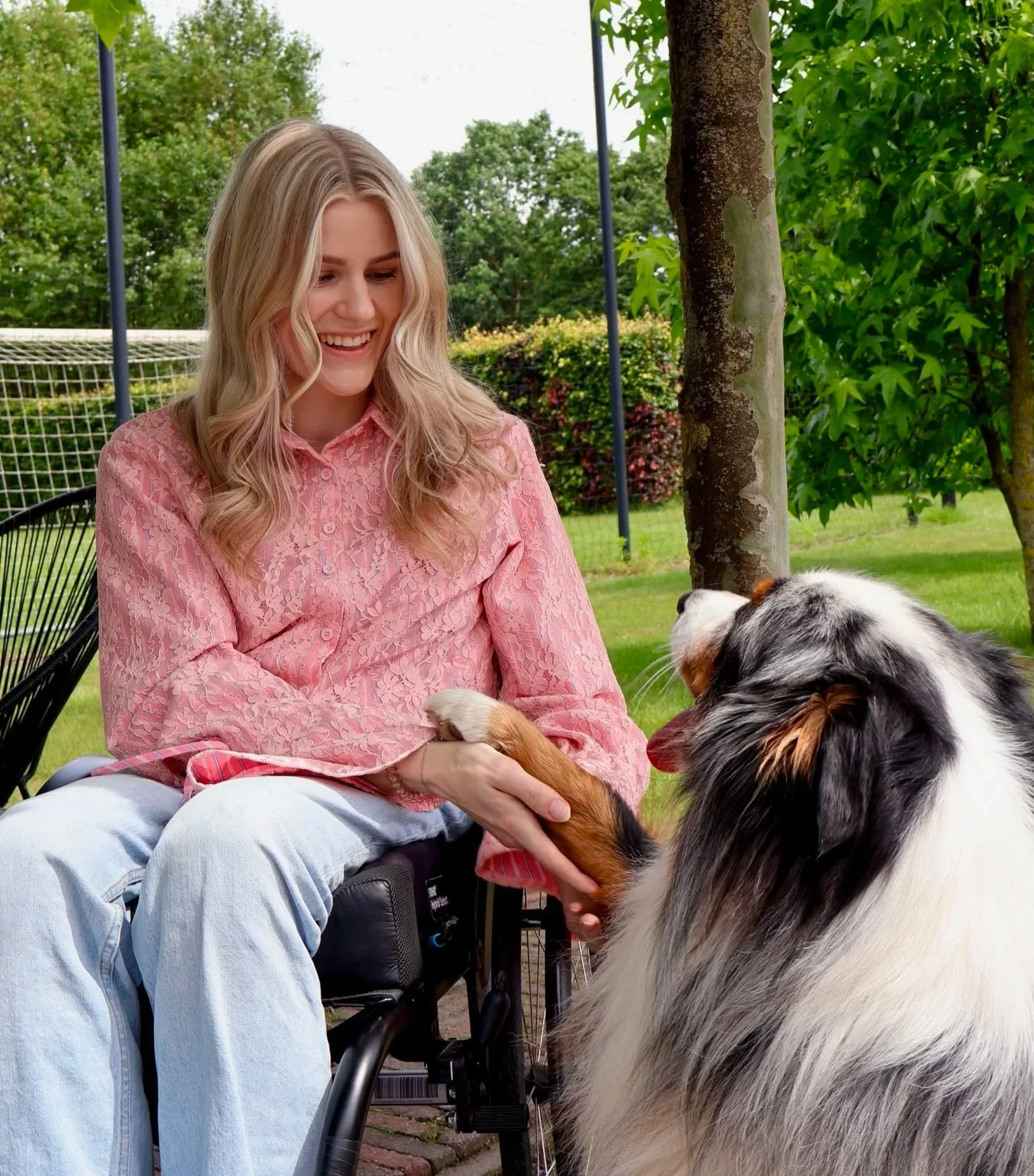 Vrouw in een rolstoel die een Border Collie aait in een tuin met groen gras en bomen.