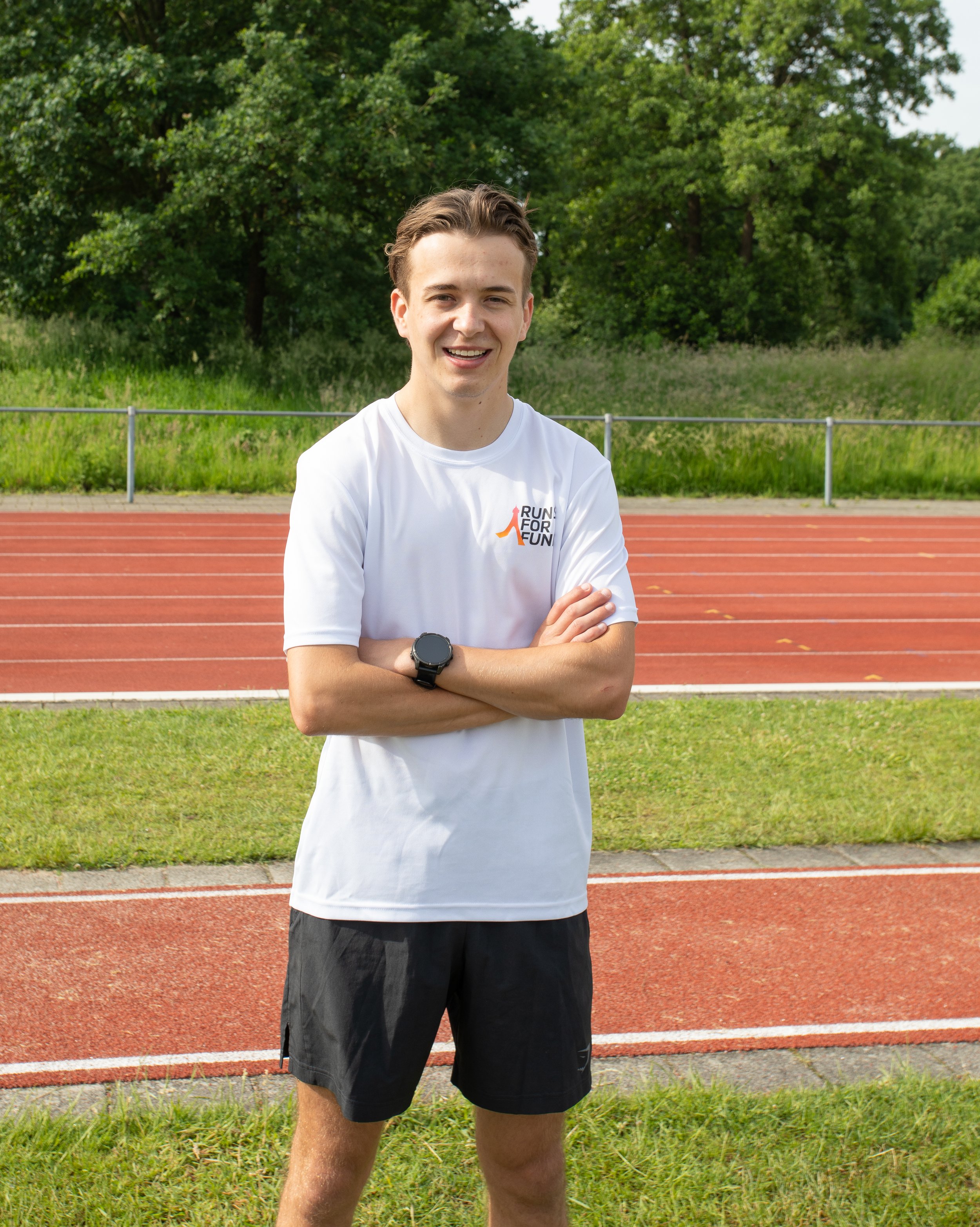 Een jonge man met een glimlach, kleurrijke sportkleding en een horloge, staat op een atletiekbaan met groene bomen op de achtergrond.