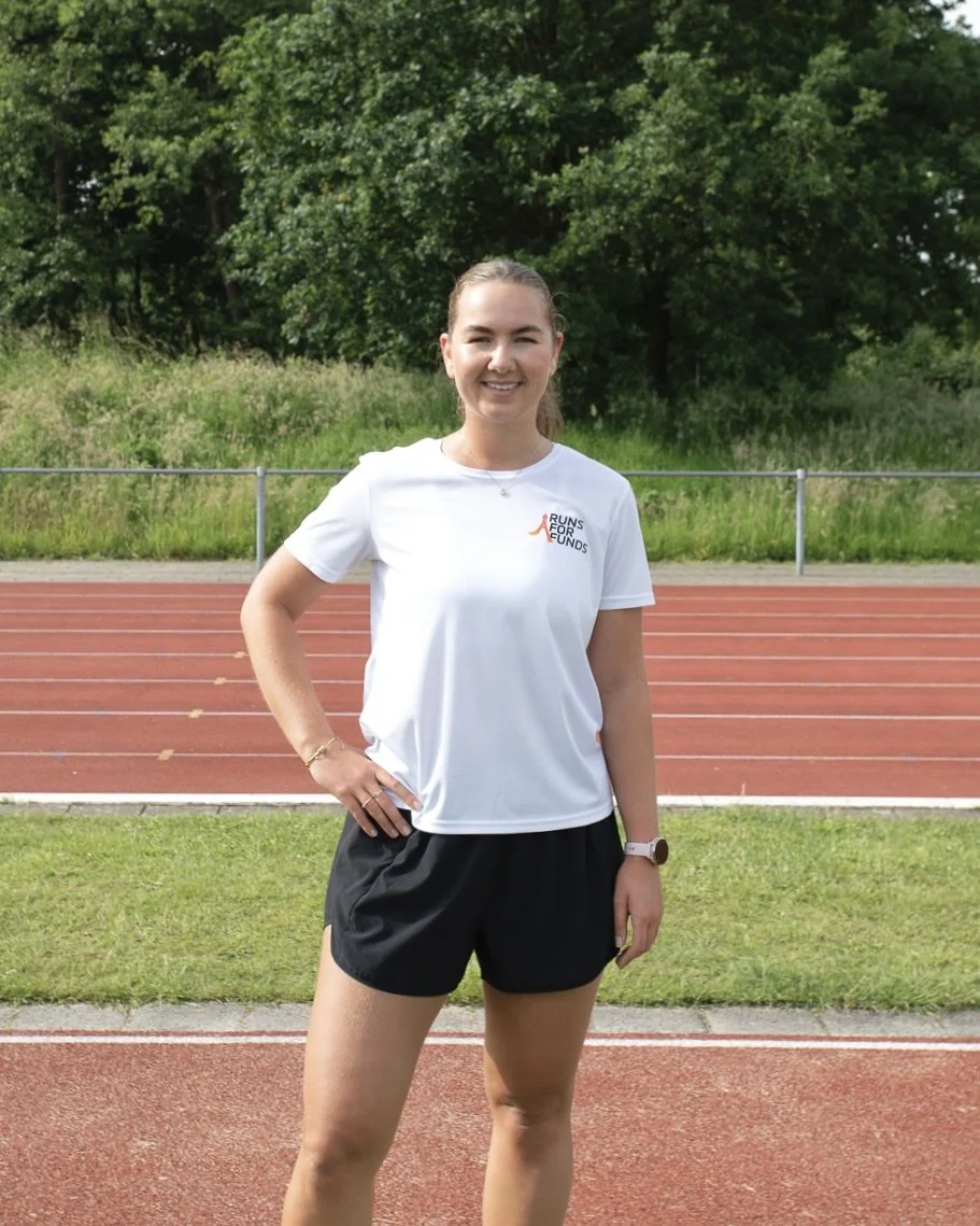 Een vrouw die op een atletiekbaan staat, gekleed in sportkleding, met een groene bos achter haar.