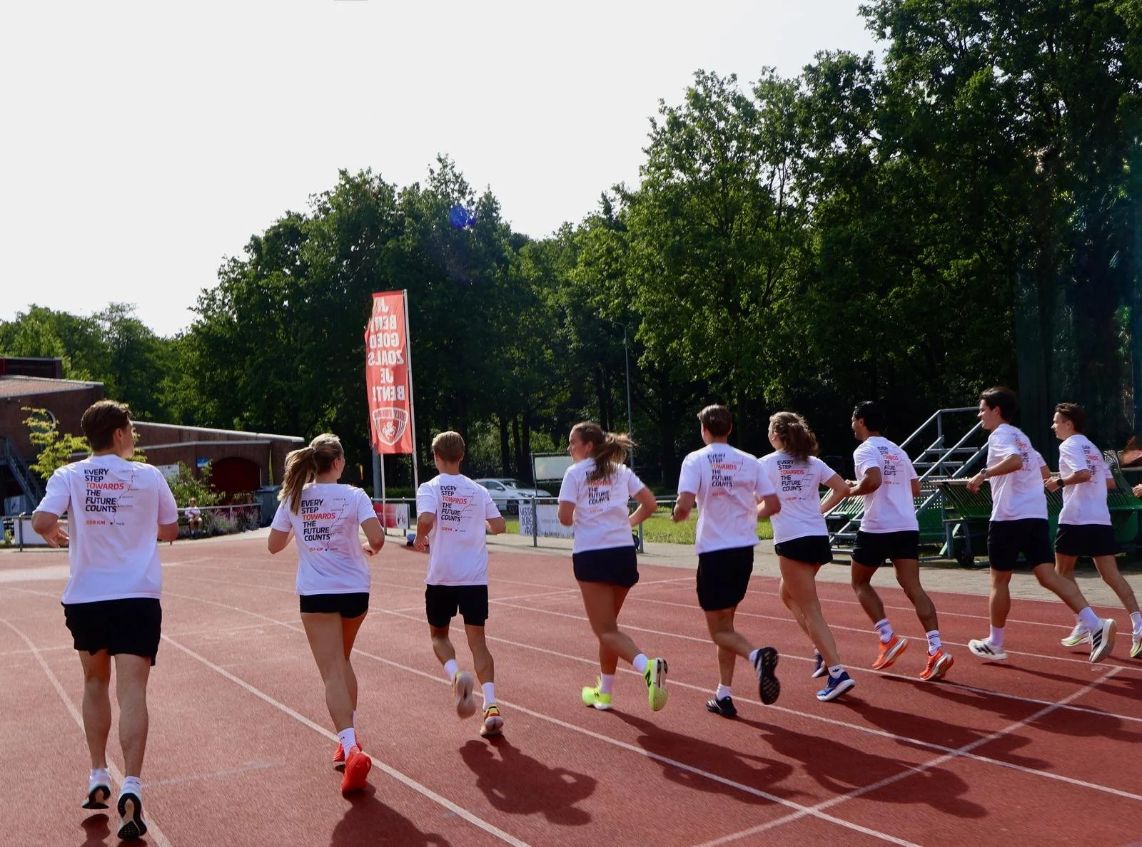 Groep lopers op een atletiekbaan tijdens een training of wedstrijd, met groene bomen op de achtergrond en een rode vlag met tekst.