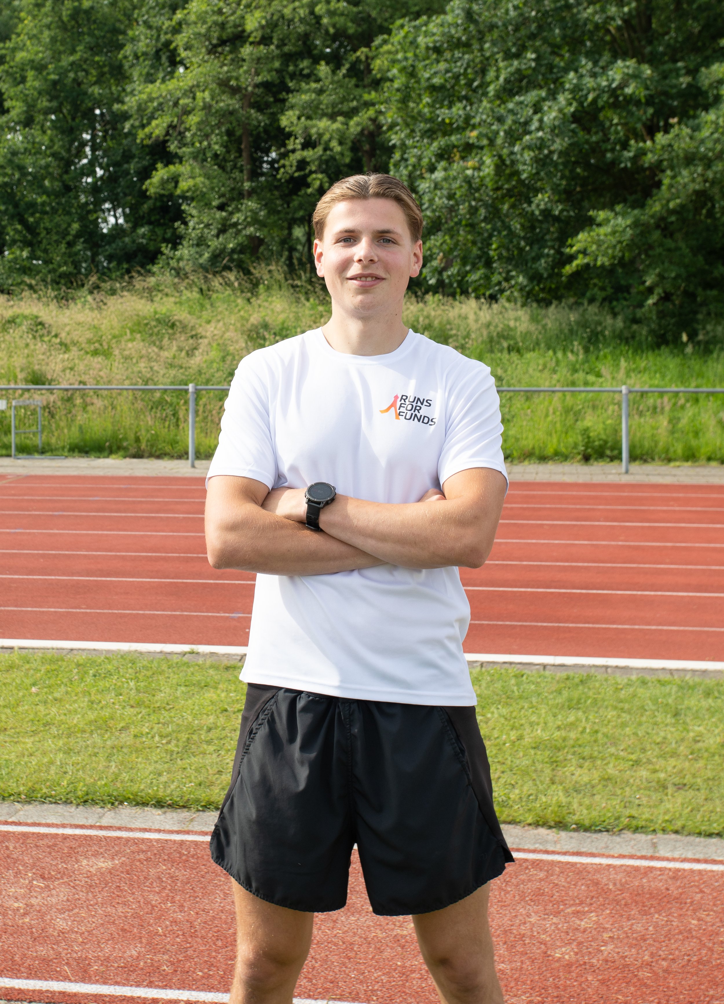 Een jonge man staat op een atletiekbaan met gekruiste armen, draagt een wit T-shirt met 'Runs for Funds' logo en zwarte shorts, met een groene bos achter hem.