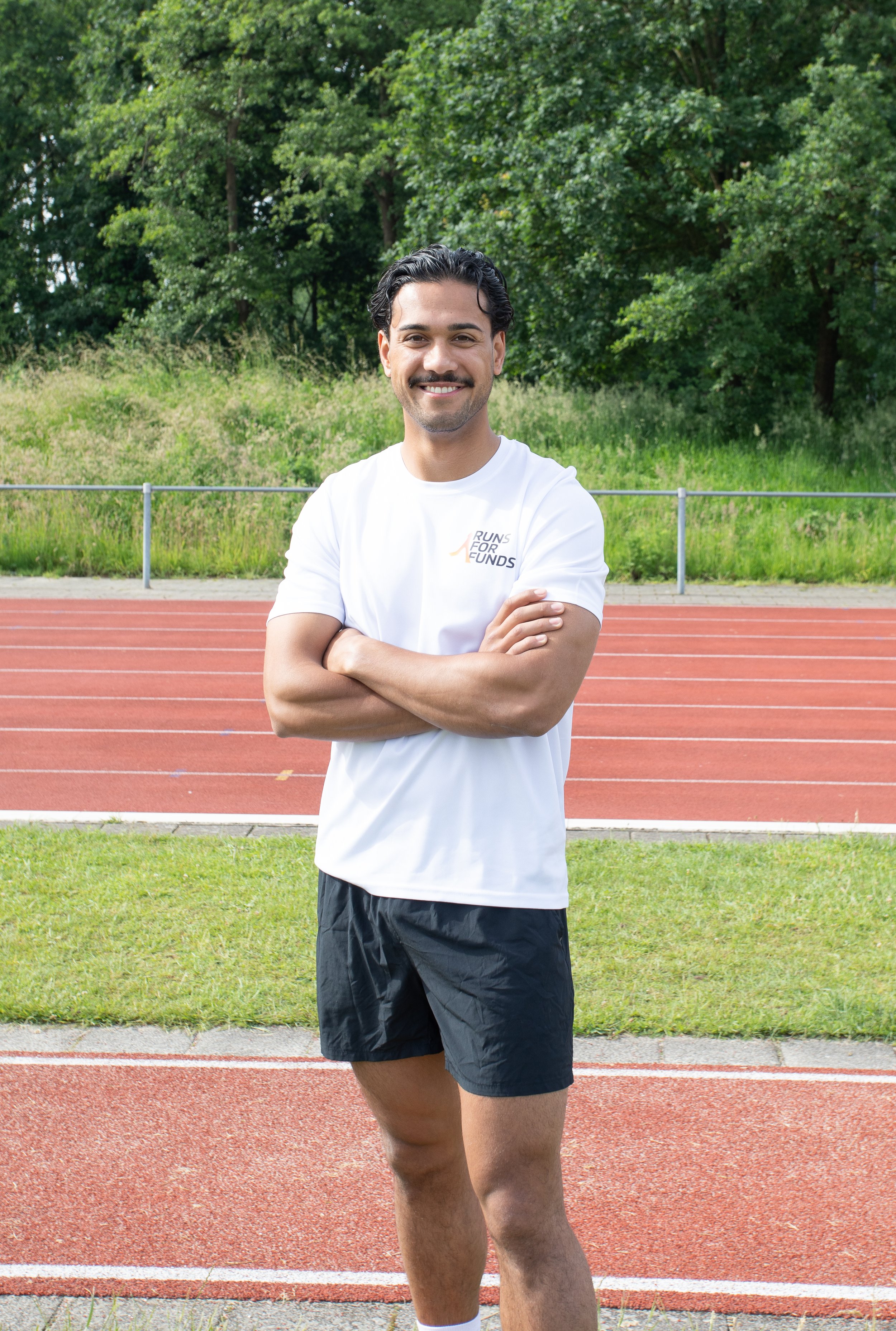 Man staat op een atletiekbaan, draagt een wit T-shirt met het logo 'RUNS FOR FUNDS' en heeft een glimlach, met groene bomen op de achtergrond.