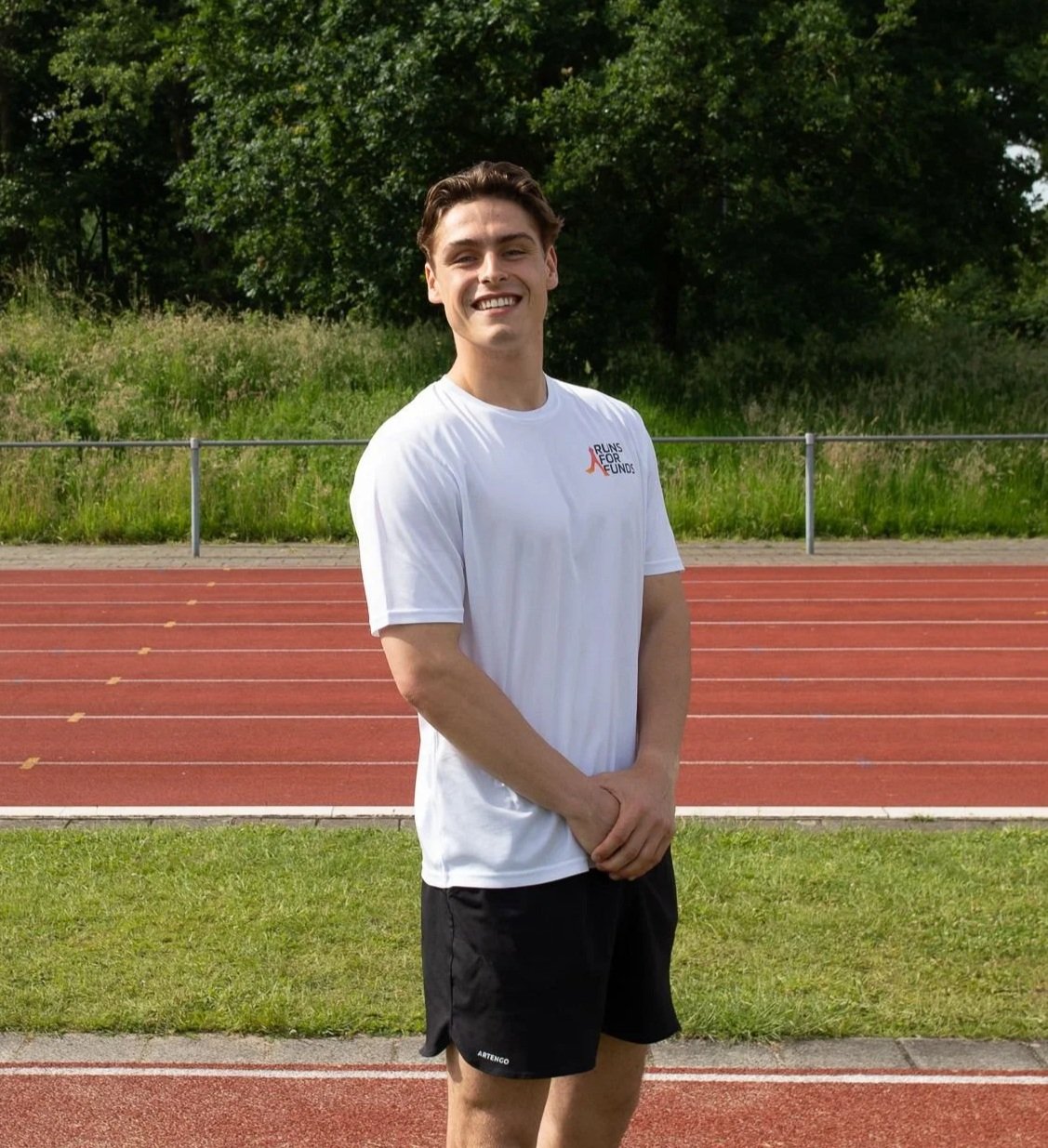 Jongen in wit T-shirt en zwarte shorts op atletiekbaan met groene achtergrond en bomen.