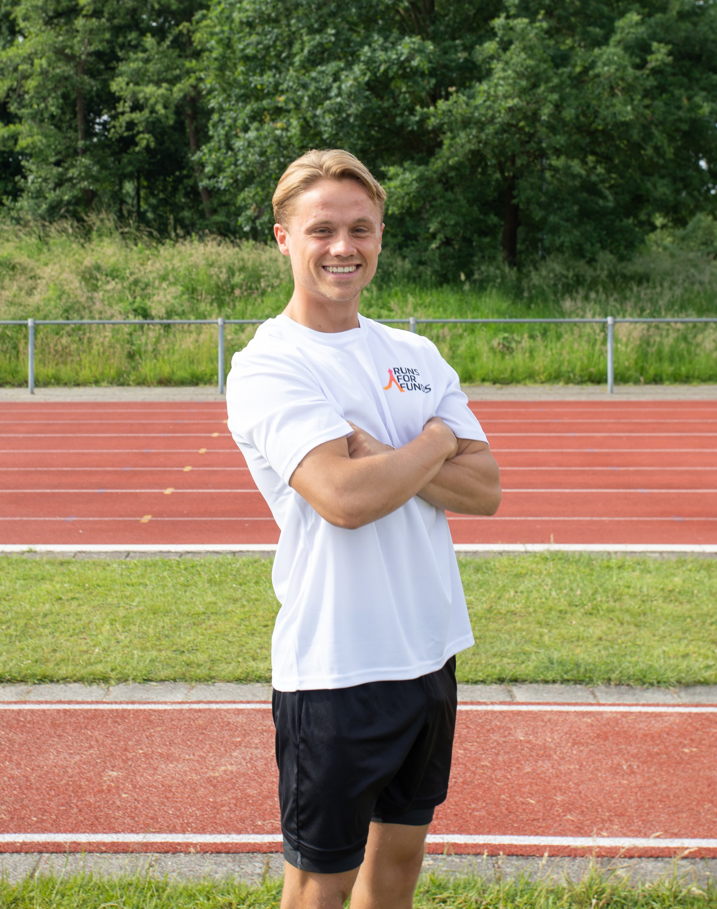 Een jonge man staat op een atletiekbaan, gekleed in een wit sportshirt met een logo en zwarte sportbroek, met zijn armen over elkaar en een glimlach, achtergrond van bomen en een hek.