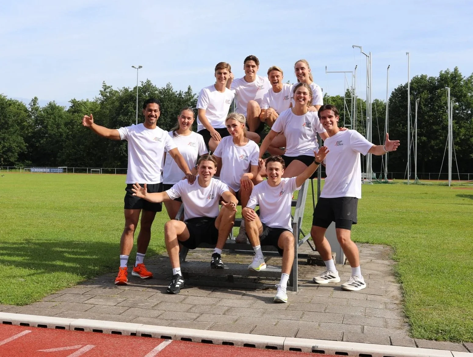 Een groep jonge atleten poseert samen op een atletiekbaan, met een groene achtergrond van bomen en een sportveld. Ze dragen witte sportkleding en lachen blij.