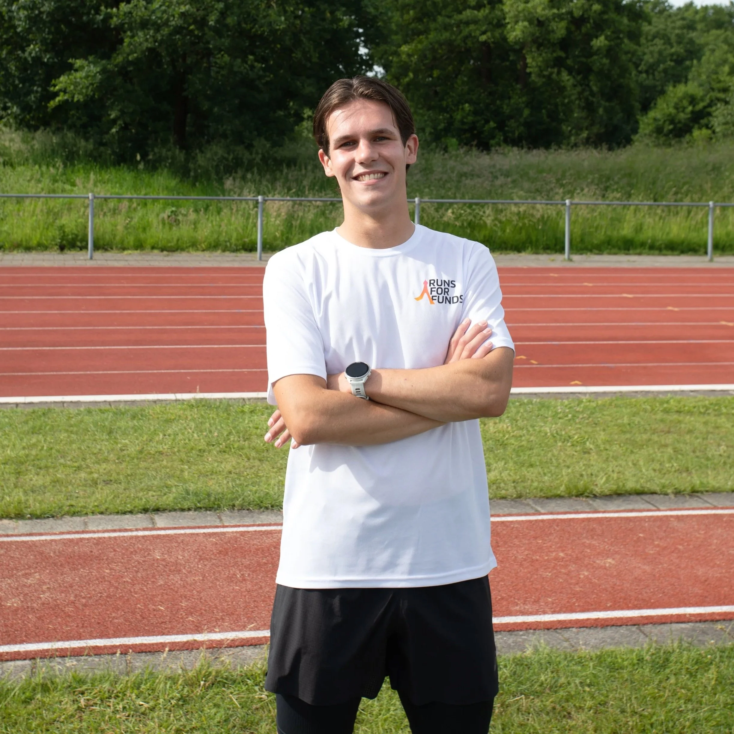 Een man met een witte sportshirt en zwarte korte broek staat op een atletiekbaan, met groene bomen op de achtergrond.
