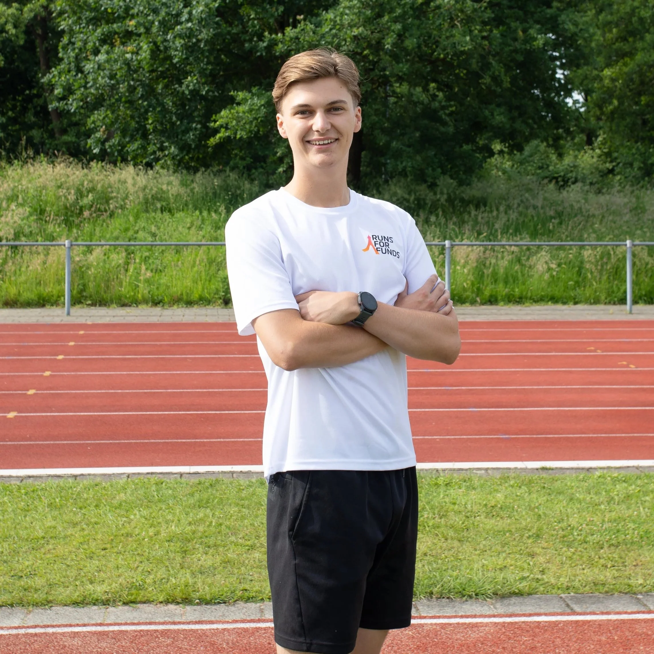 Een jonge man met bruin haar, glimlachend en gekleed in een wit T-shirt met het logo 'Runs for Funds', staat met gekruiste armen op een atletiekbaan, met groene bomen op de achtergrond.