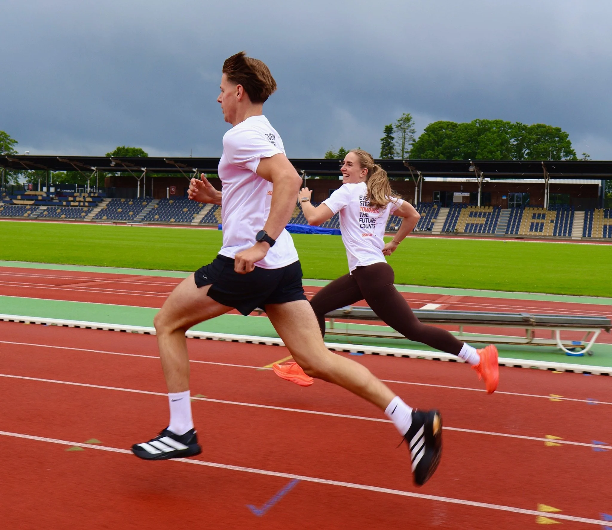 Twee vrouwen rennen op een atletiekbaan op een bewolkte dag, met lege tribunes en groene bomen op de achtergrond.