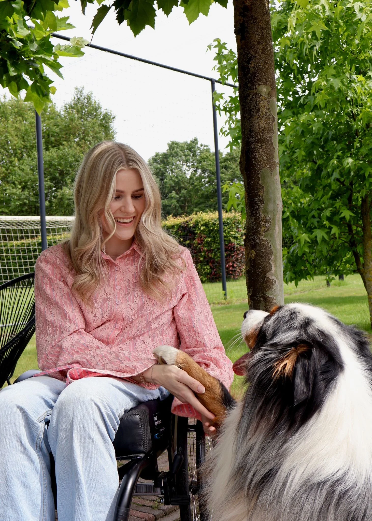 Een jonge vrouw met lang blond haar glimlacht terwijl ze haar hand uitstrekt naar een Australian Shepherd-hond die haar poot vasthoudt. Ze zitten buiten in een groene tuin met bomen en een volleybalnet op de achtergrond.