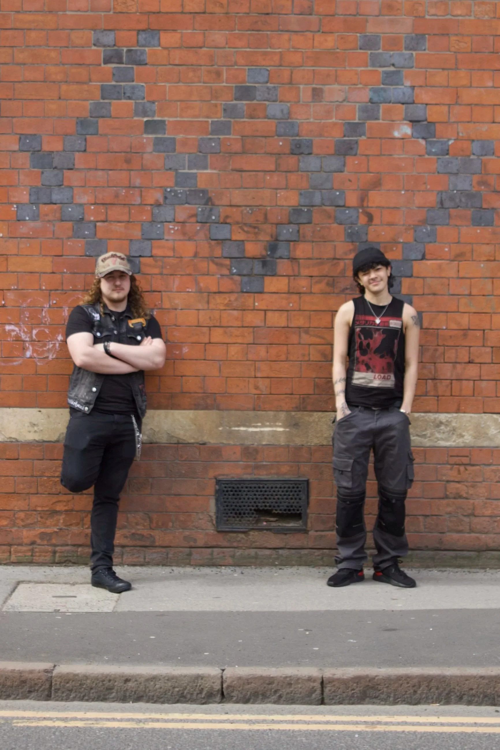 Two young men standing in front of a brick wall with a large heart shape made of black and gray bricks.