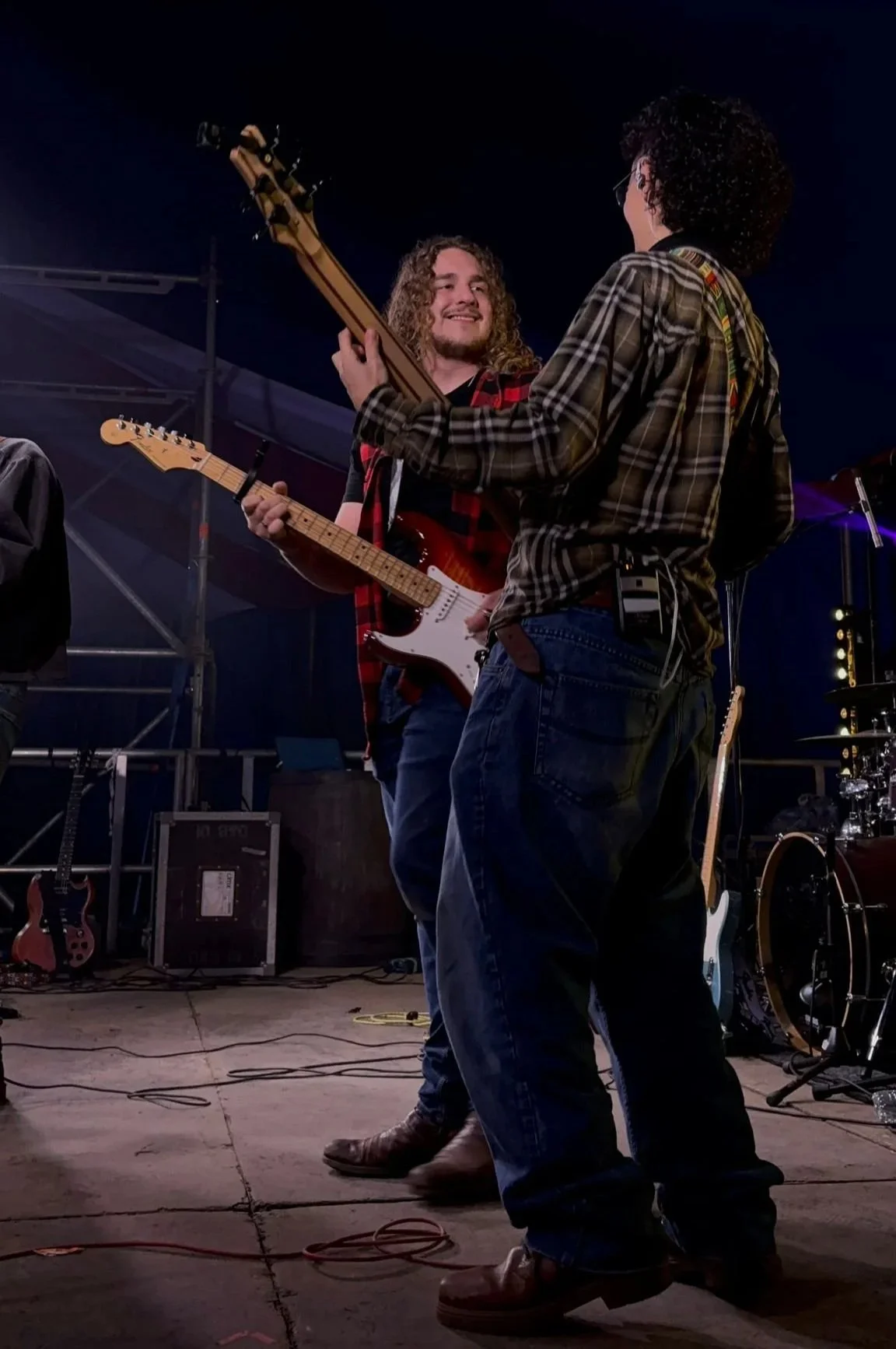 Two men holding electric guitars on stage during a live music performance, with musical equipment and instruments around them at night.