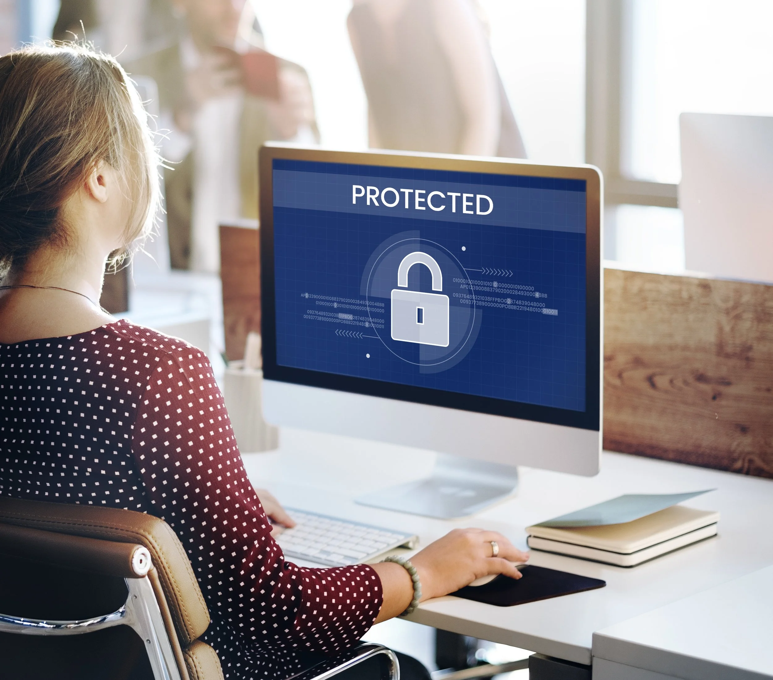 A woman with blonde hair wearing a red polka dot shirt sitting at a desk in front of a computer with a blue screen displaying the word 'PROTECTED' and a lock icon, in a well-lit office environment.