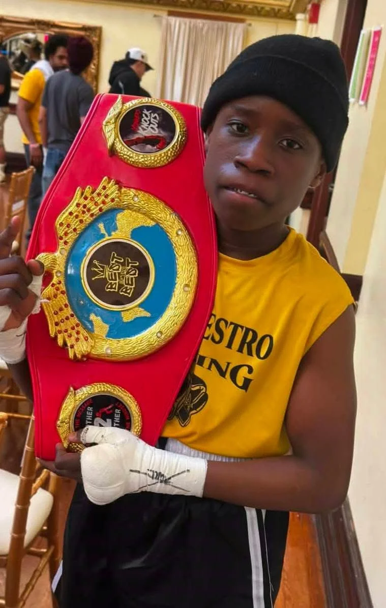 Young boy in a yellow sleeveless shirt holding a red boxing championship belt, wearing a black beanie and boxing wraps on one hand, in a room with other people and chairs.
