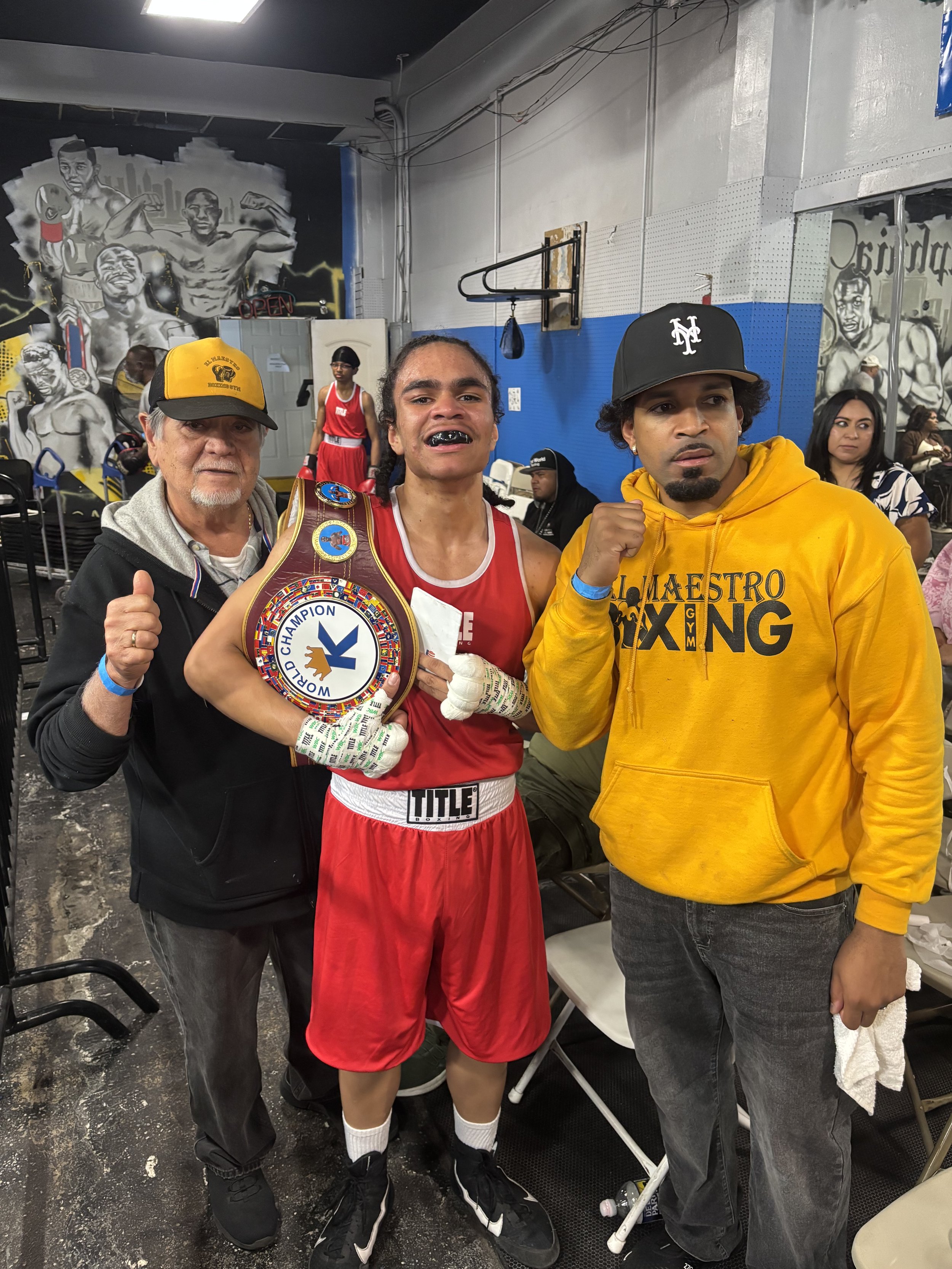 A male boxer in red shorts and gloves is holding a championship belt, flanked by two men, one in a black jacket and yellow cap and the other in a yellow hoodie, with a boxing gym and mural in the background.