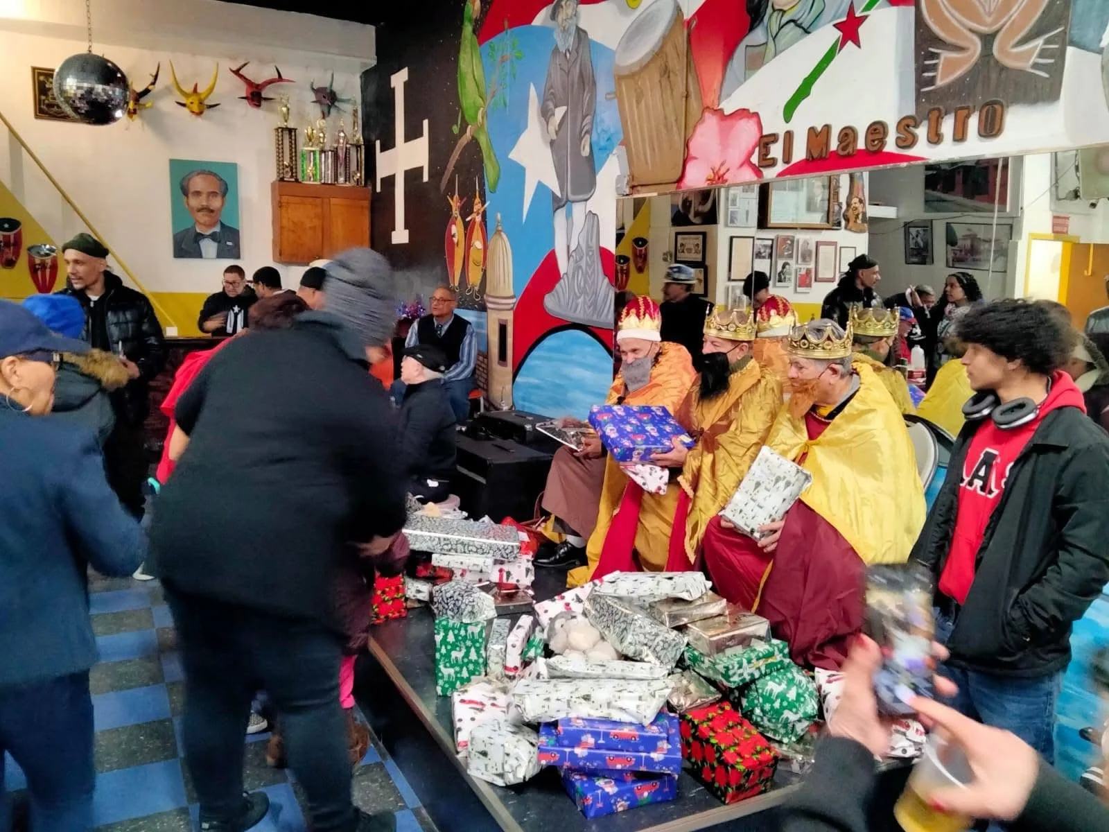 People gathered in a decorated room with a mural of a man in a suit and a large Panama flag. Some attendees are dressed as kings with crowns and robes, exchanging gifts on a table filled with wrapped presents. The scene appears festive, with a birthday cake and party decorations.