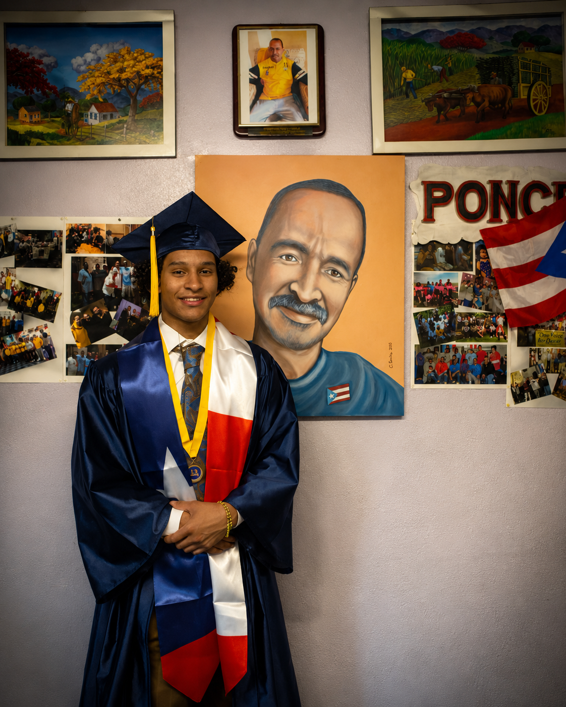 A young man in a graduation gown and cap standing in front of a colorful mural with portraits and photos, smiling at the camera.