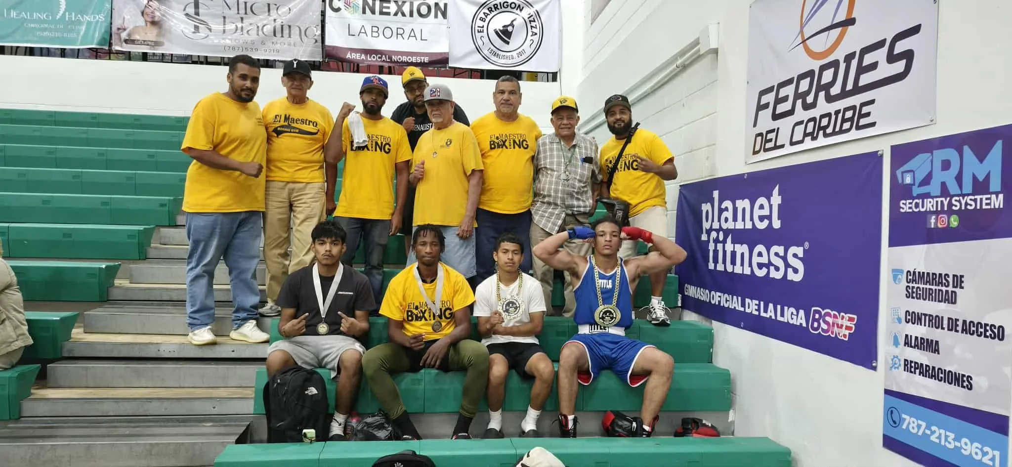 A group of people, including young martial artists with medals and coaches, poses on green bleacher seats inside a gymnasium after a boxing competition. The then coaches and trainers are standing behind the young athletes, some of whom are sitting or standing with medals, in front of banners advertising fitness and security services.