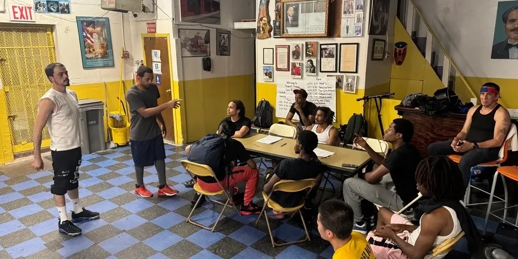 A diverse group of young people gathered in a casual, indoor community space, with some seated around a table and others standing. The room has yellow and gray walls, various posters, photographs, and artworks, with stairs leading to an upper level. A person standing appears to be speaking or leading a discussion, while others listen attentively.