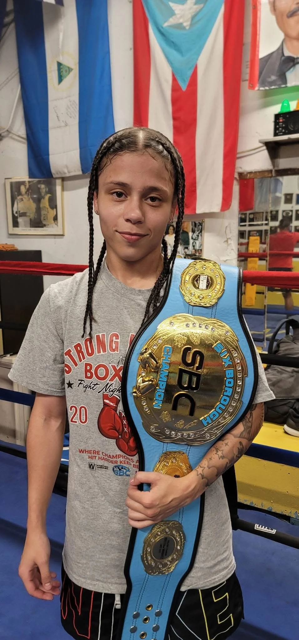 A young female boxer with braided hair holding a large championship belt in a gym, with boxing posters and flags in the background.