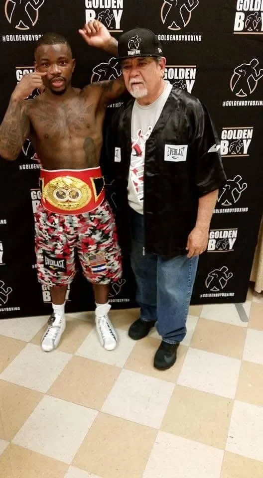 A young shirtless male boxer with a championship belt around his waist, wearing patterned shorts and white boxing shoes, standing next to an older man in a black jacket, baseball cap, and jeans, both posing with fists raised in front of a backdrop with boxing branding.