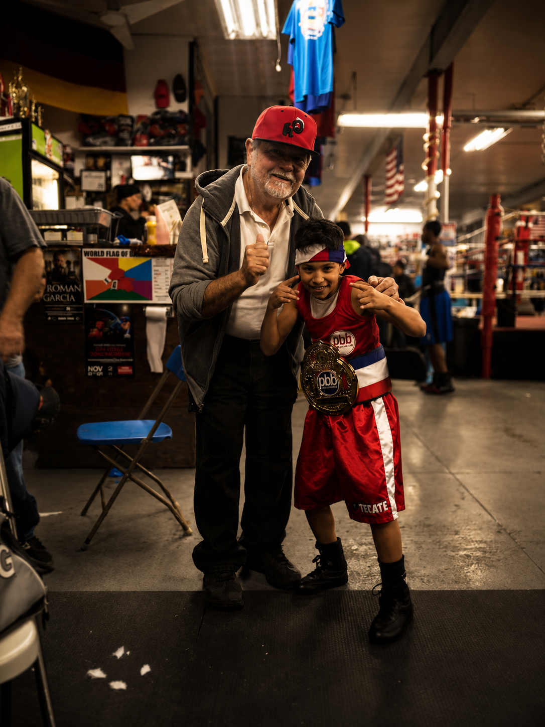 A young boy in boxing gear with a championship belt and a man with gray hair and a beard, both smiling and posing together inside a gym or boxing gym.