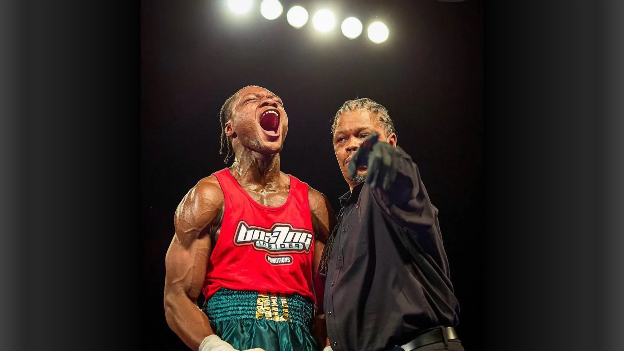 A muscular boxer in red trunks with gloves raising his head and yelling, standing beside a man in a black shirt pointing at the boxer, against a dark background with bright overhead lights.
