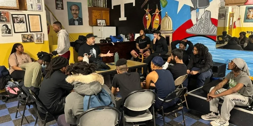 A group of people sitting around a table in a colorful room, listening to a man in a black hat and shirt speaking. The room has yellow and black walls with photographs, paintings, and a mural depicting a puerto rican theme.