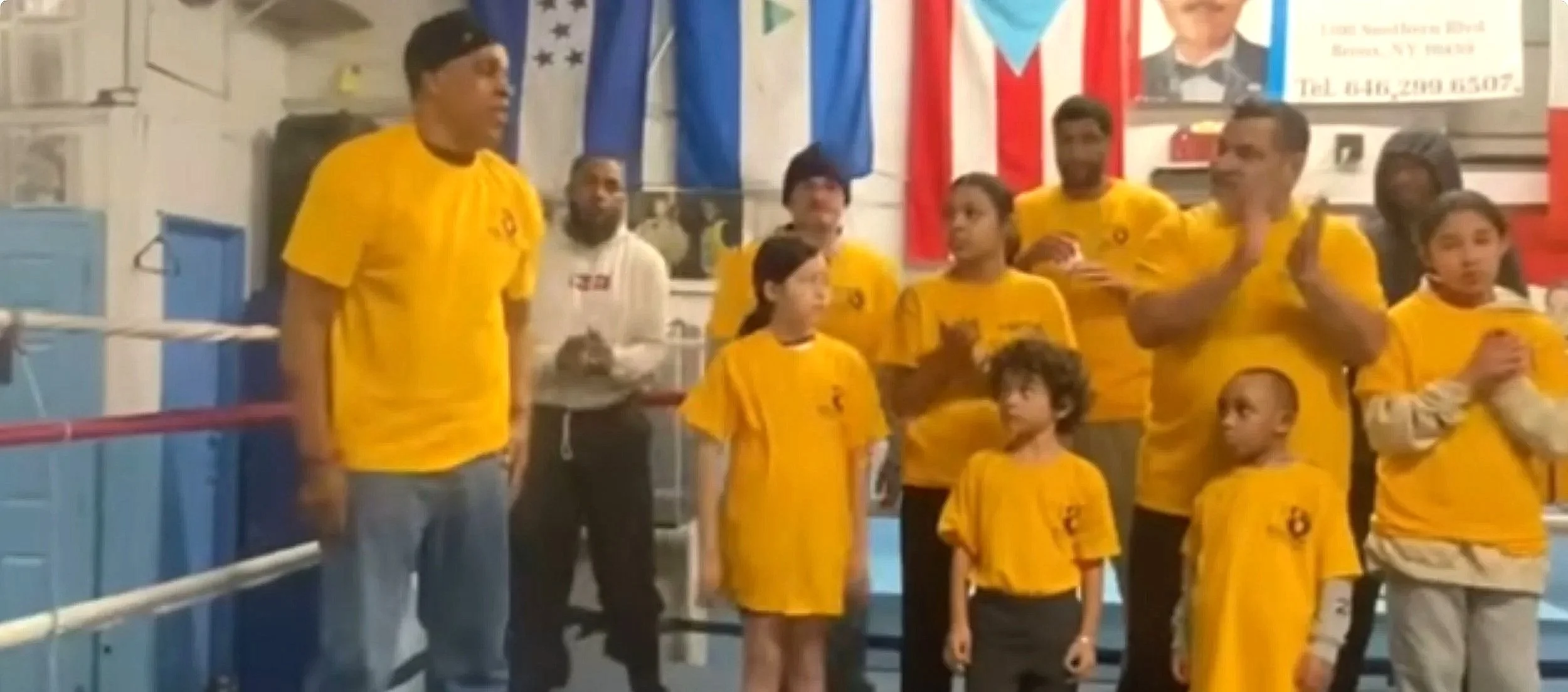A group of people, including children and adults, wearing yellow shirts, standing inside a boxing gym with a boxing ring and flags in the background.