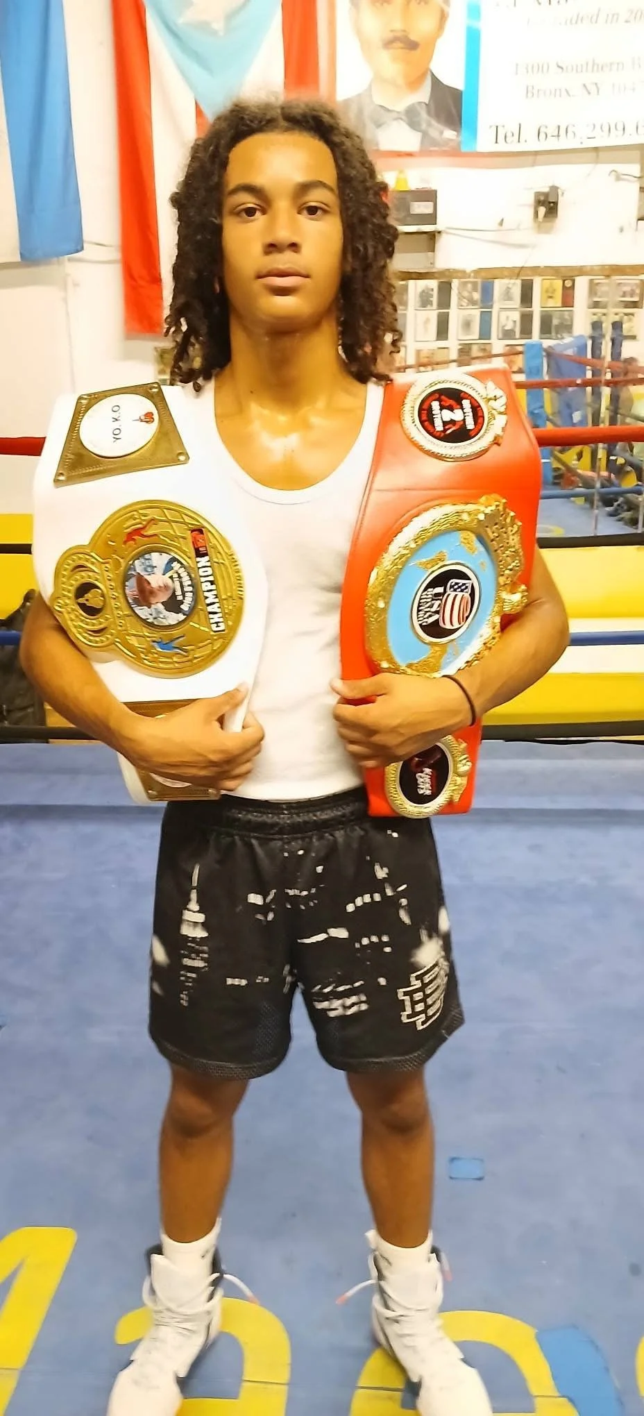 A young male boxer standing in a boxing gym, holding two championship belts across his chest. He is wearing a white t-shirt, black shorts, and boxing shoes.