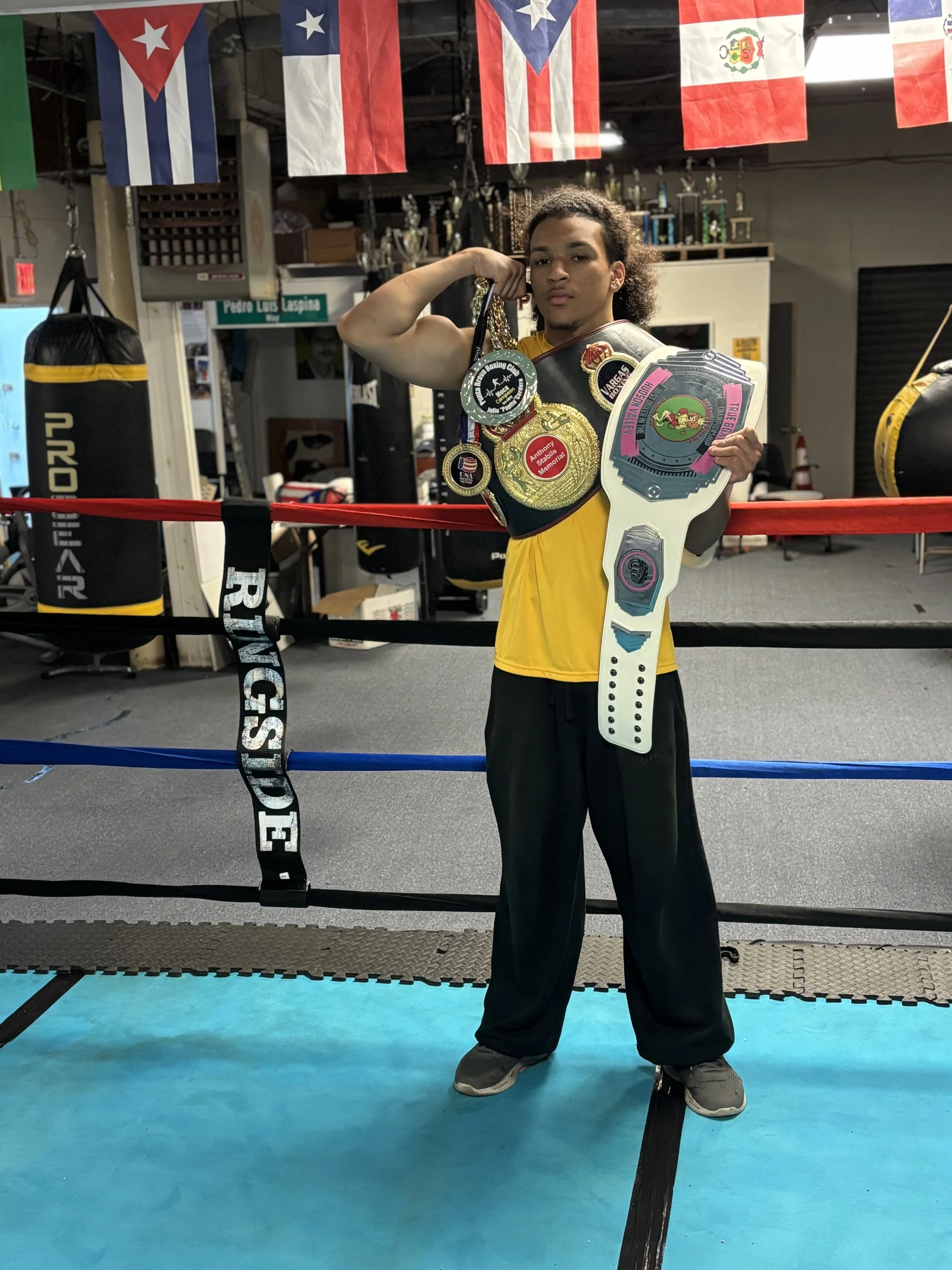 A young man with curly hair standing in a boxing gym, flexing his right arm with multiple championship medals hanging from his neck, and holding a cardboard cutout of a championship belt in his left hand. The gym has boxing rings, punching bags, and flags from various countries hanging from the ceiling.