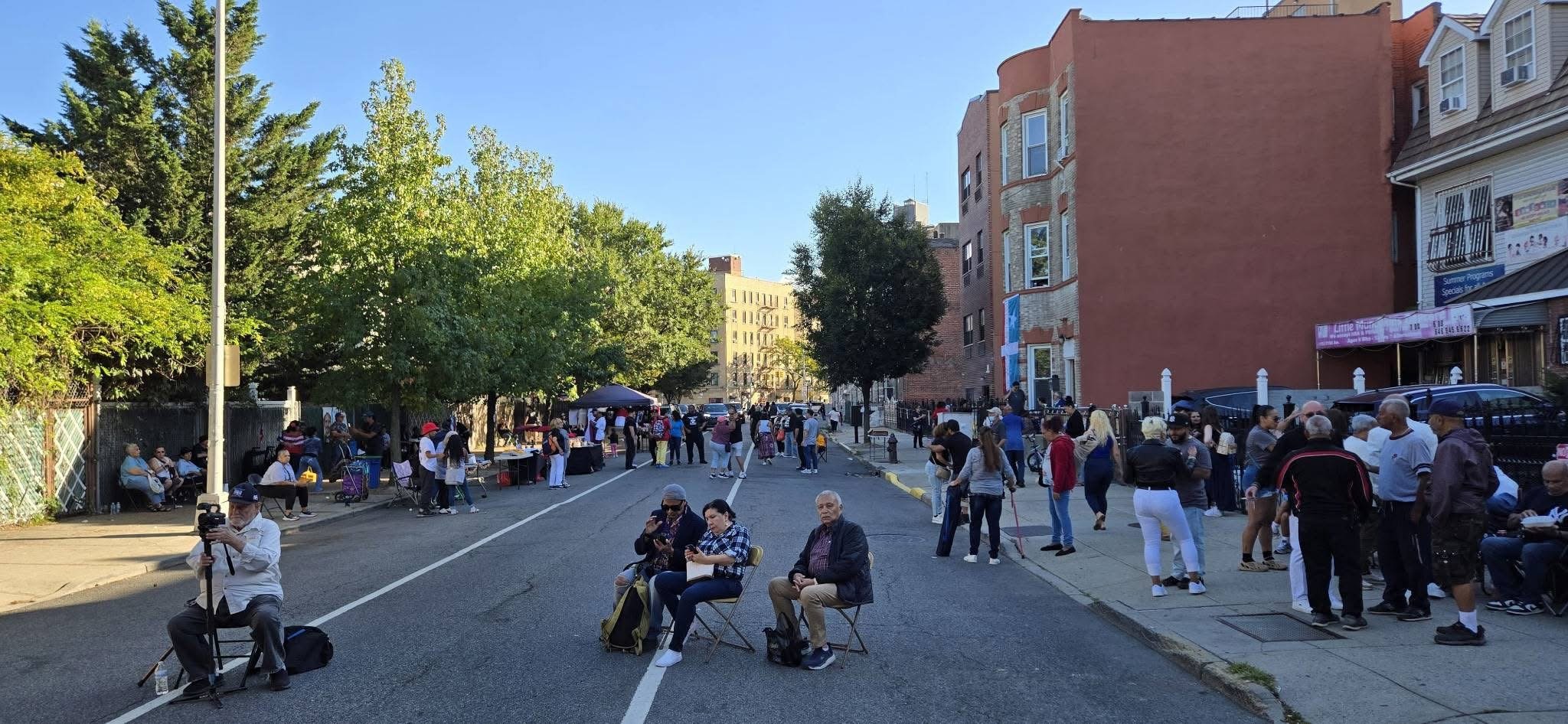 A street scene during a community event with people sitting and standing, vendor tables, and a small crowd gathered on a sunny day in an urban area with trees, buildings, and a clear blue sky.