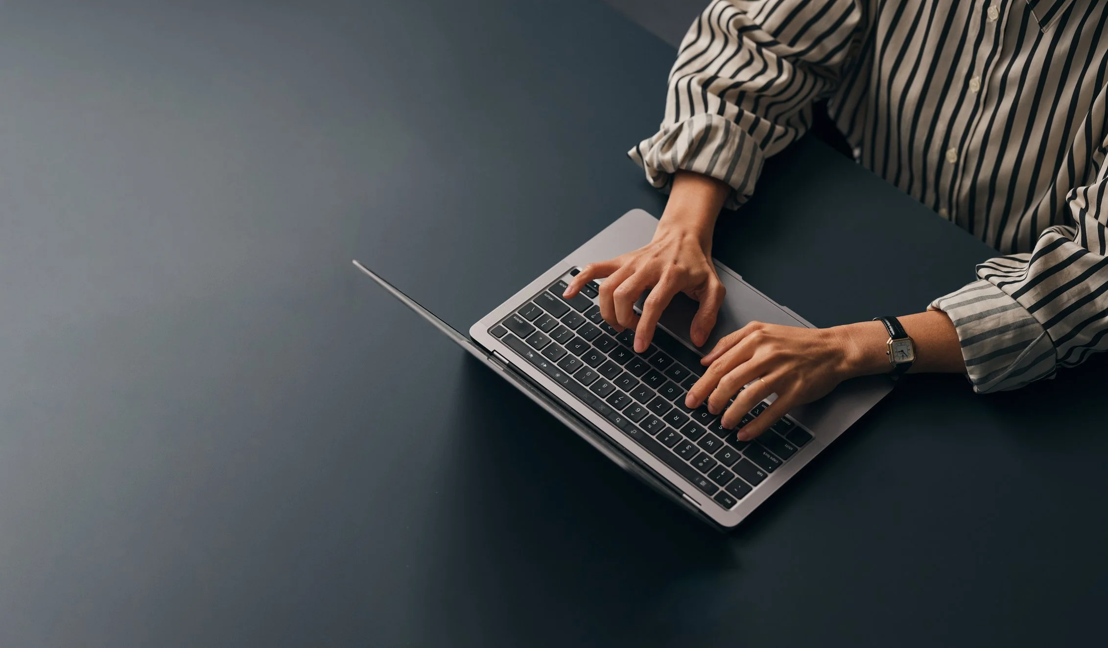 Person typing on a silver laptop with a striped shirt and watch, on a dark table.