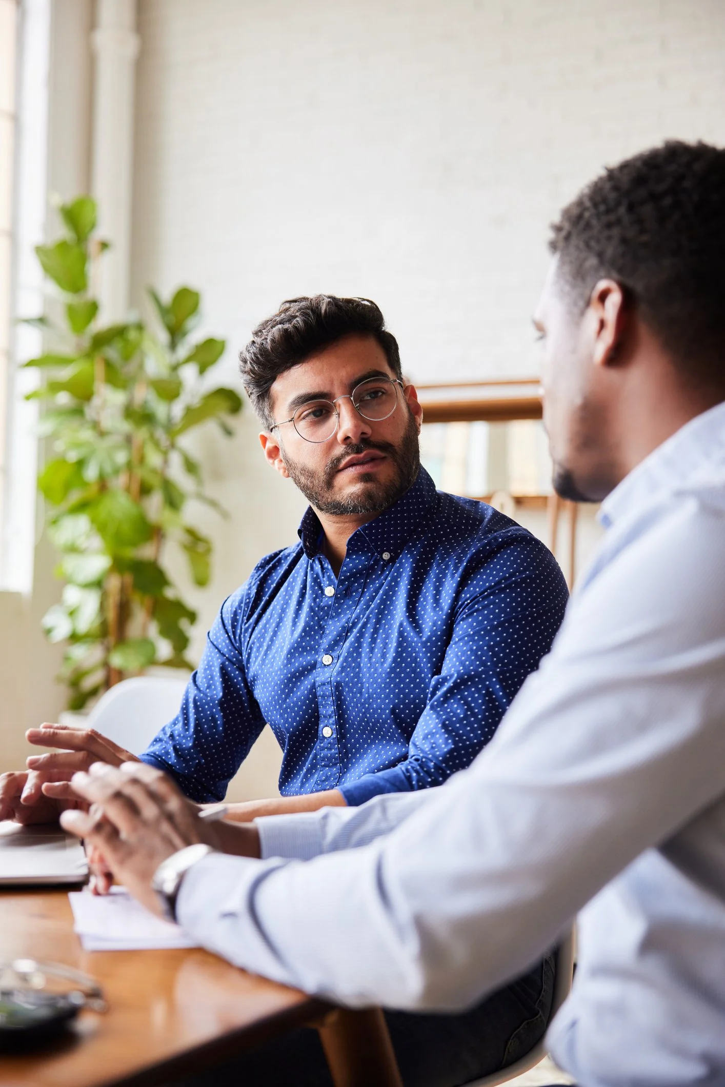 Two men are sitting at a table engaged in conversation. One man has glasses, a beard, and a blue polka-dotted shirt, while the other has short, curly hair and a white shirt. There are notebooks and a plant in the background.