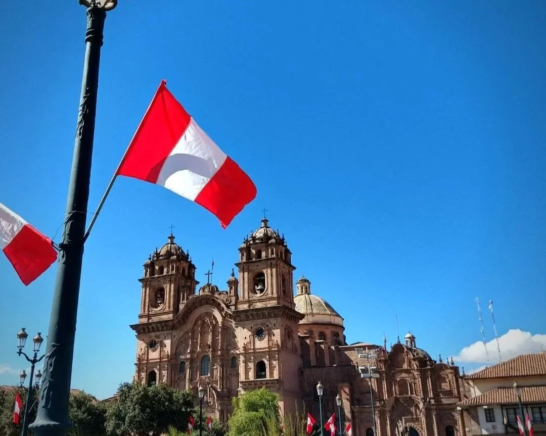 Catedral en Cusco con banderas de Perú en primer plano y cielo azul de fondo.
