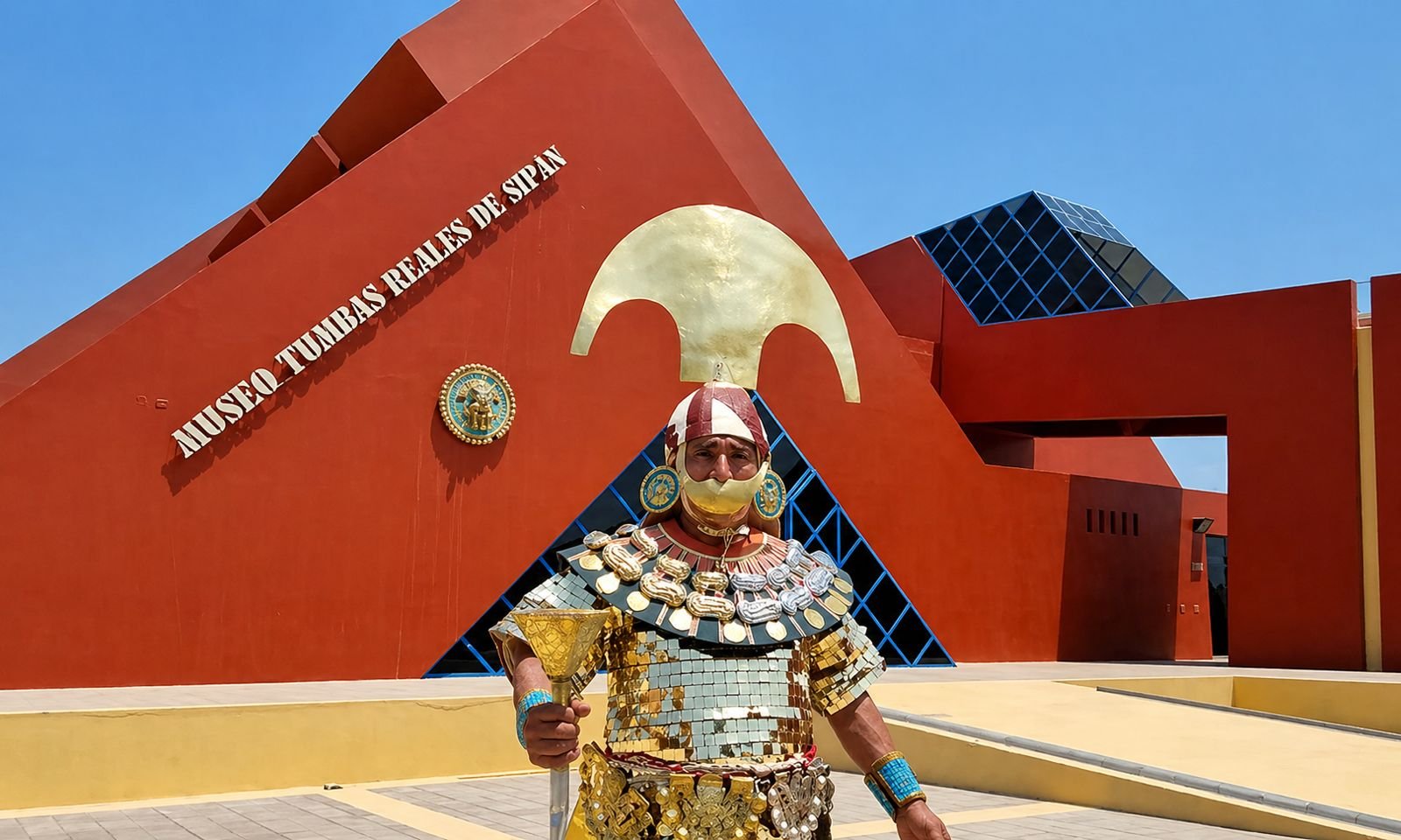 Una persona vestida con ropa y accesorios de oro, con un tocado y máscara, posando en frente del Museo Tumbas Reales de Sipán, un edificio moderno de color rojo con formas geométricas y un arco dorado en la fachada.