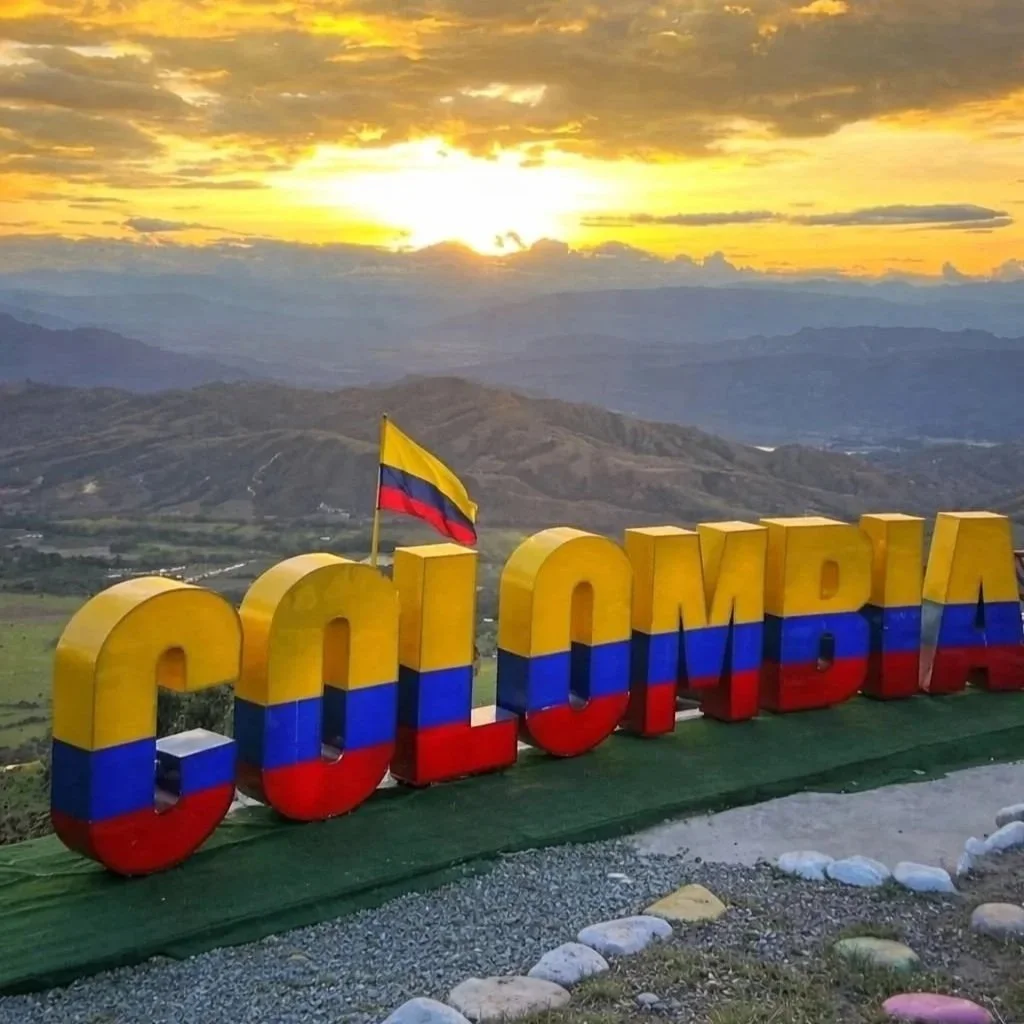 Vista de un cartel grande que dice "Colombia" pintado con los colores de la bandera nacional, con una bandera colombiana ondeando en la parte superior, al atardecer con montaña y un cielo nublado y dorado al fondo.