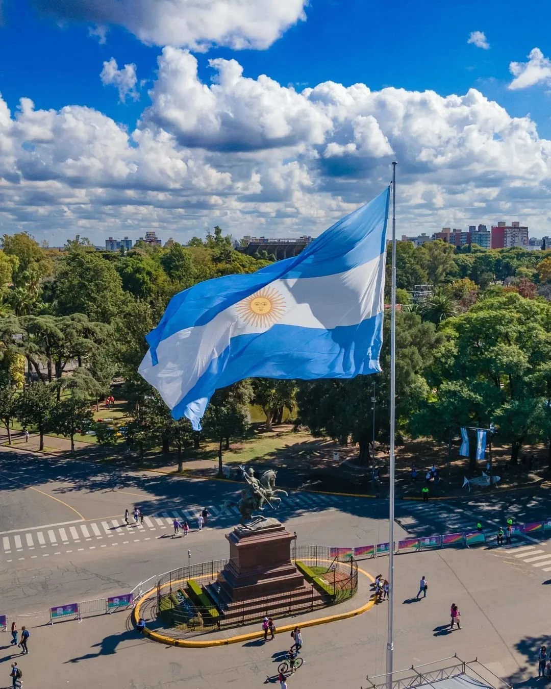 Bandera de Argentina ondeando en un parque bajo un cielo con nubes, con una estatua ecuestre y árboles verdes en el fondo.