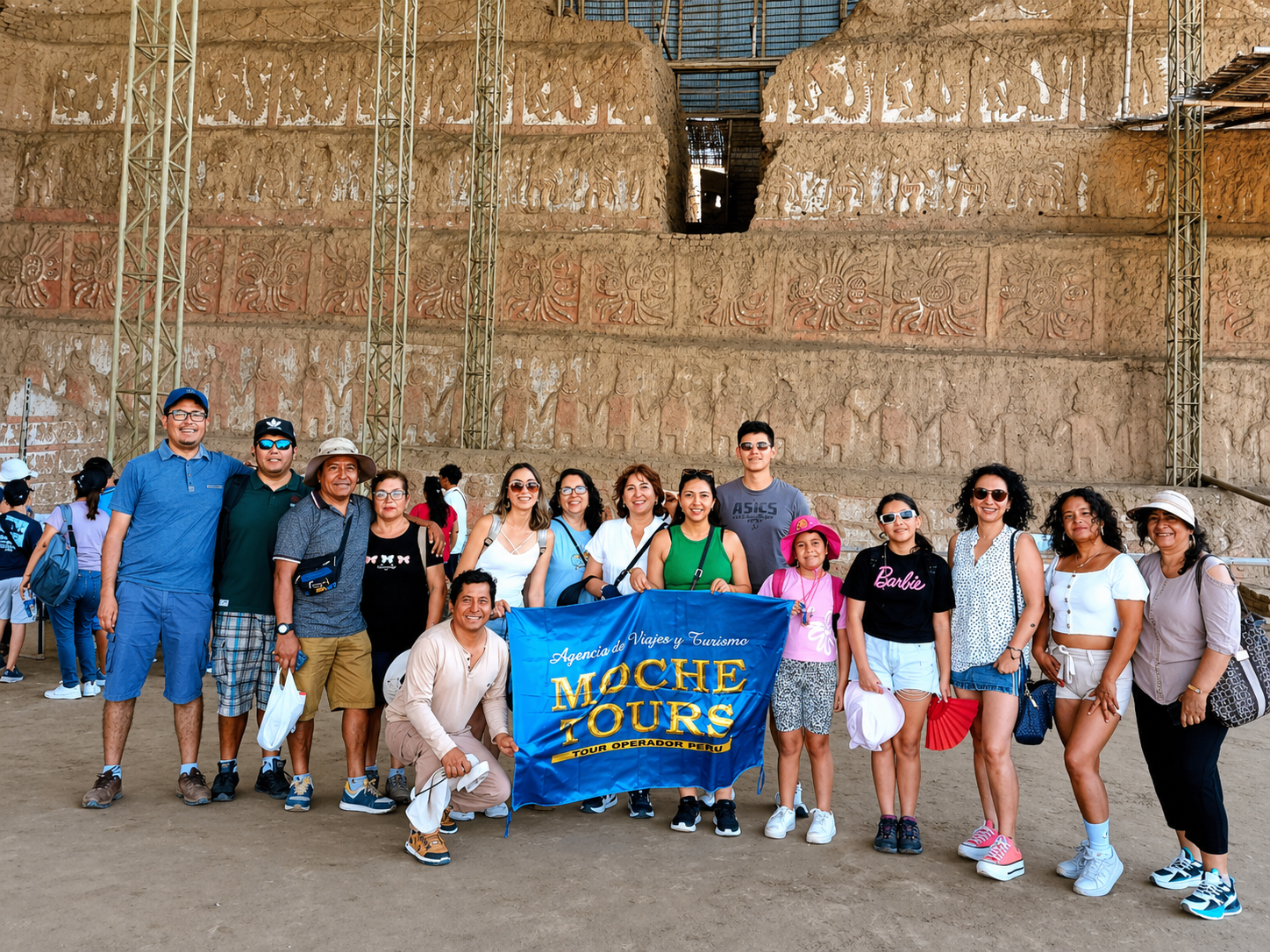 Grupo de turistas sonriendo con un cartel que dice 'Moche Tours' frente a un muro de cerámica de la cultura Moche en Perú.