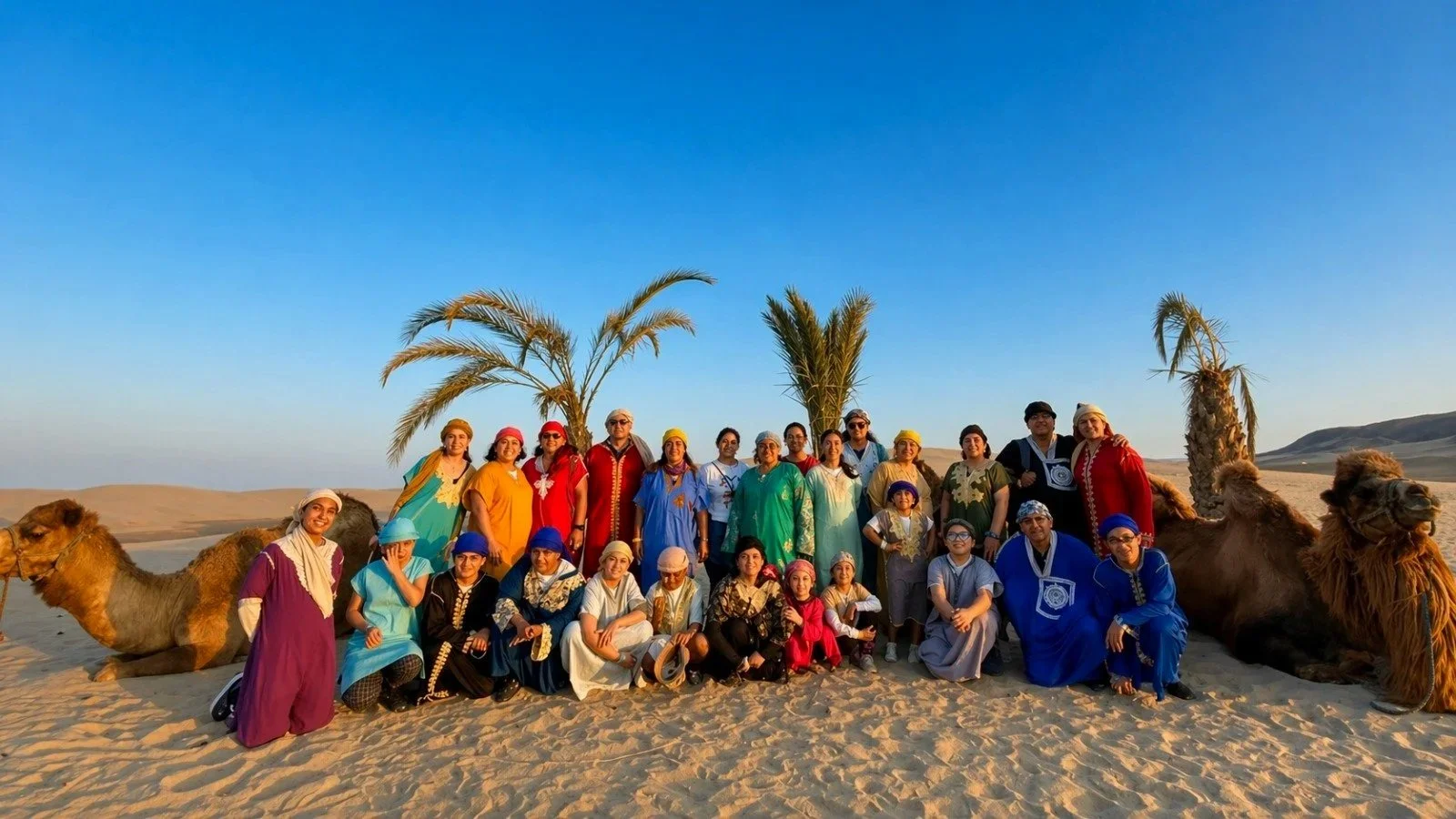 Grupo de personas vestidas con ropa tradicional en un desierto, con palmeras y camellos al fondo.