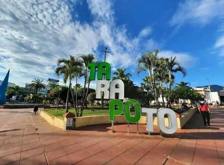 Cartel con letras grandes que dicen "TARAPOTO" en una plaza con palmeras, cielo azul y pocas personas.