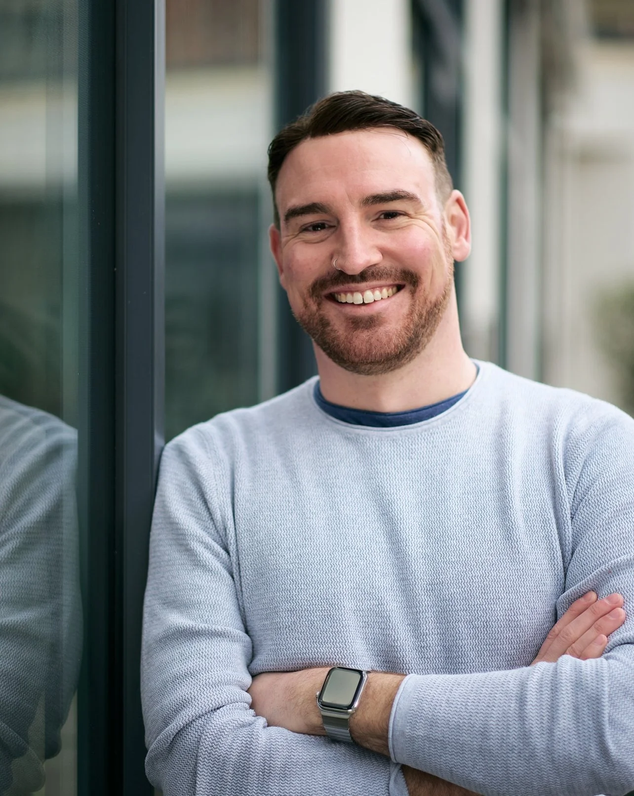 A smiling man with dark hair and a beard, wearing a light gray sweater with a navy shirt underneath, standing with arms crossed near a glass wall.