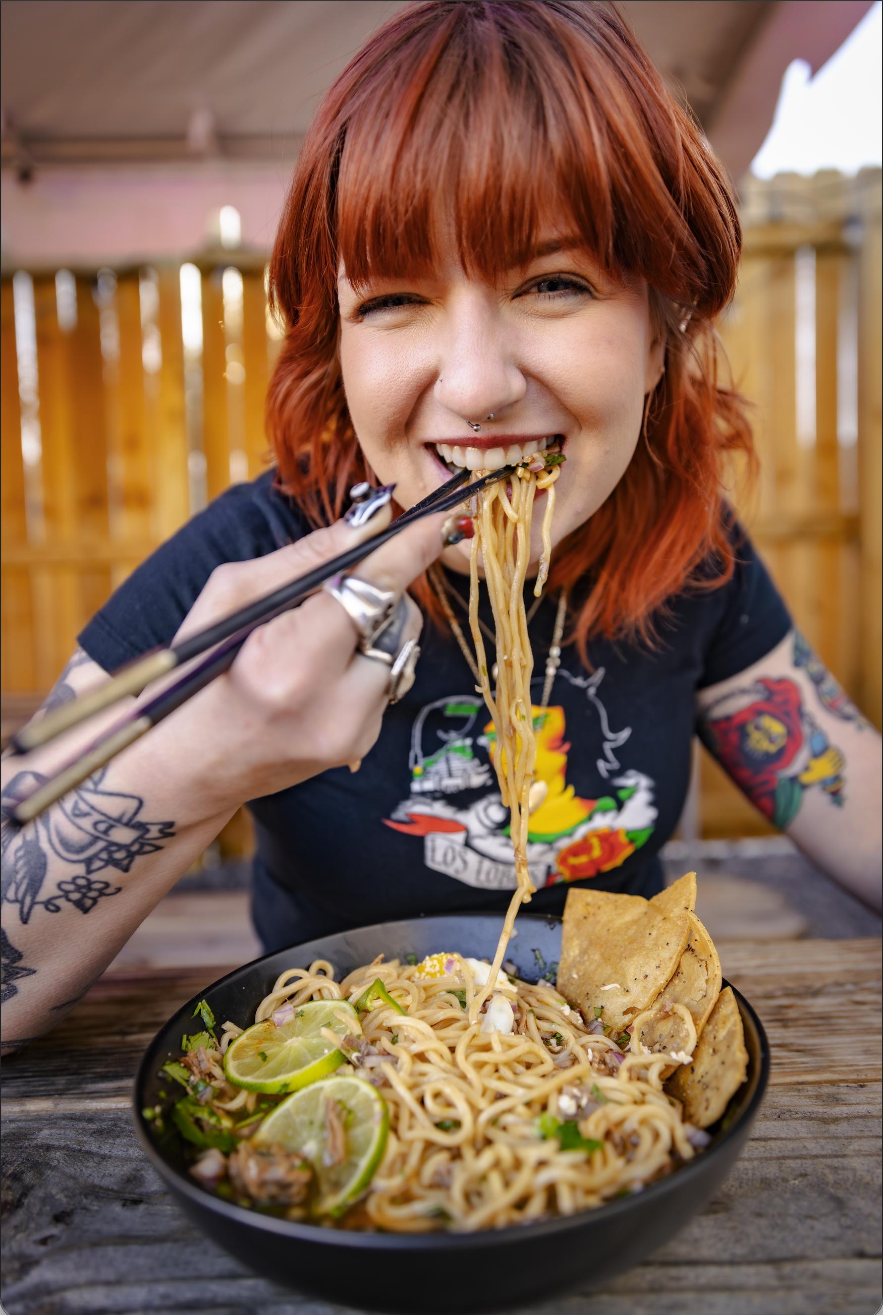 A young woman with red hair and tattoos on her arm is eating a bowl of ramen noodles with chopsticks. The bowl contains lime wedges, vegetables, and slices of meat, set on a wooden table outside.