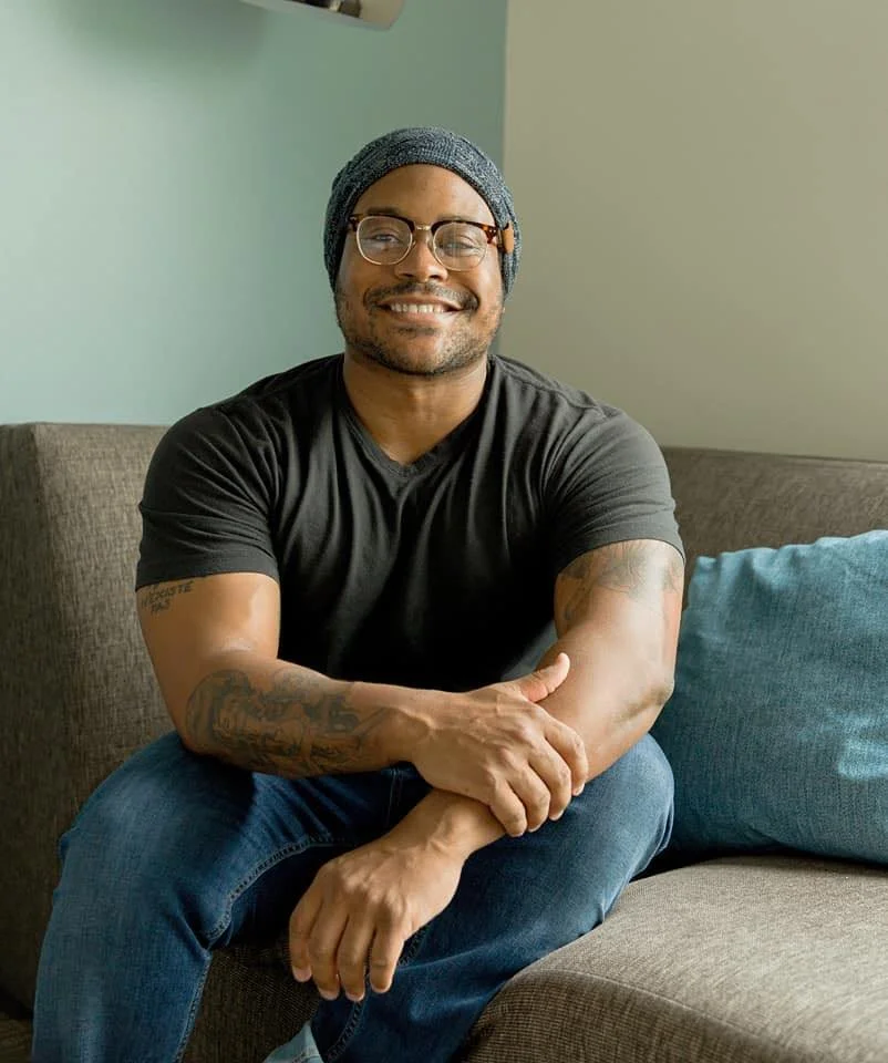 A smiling man with glasses and a beanie sitting on a sofa with his arms crossed, in a cozy room with a light green wall and a blue pillow.