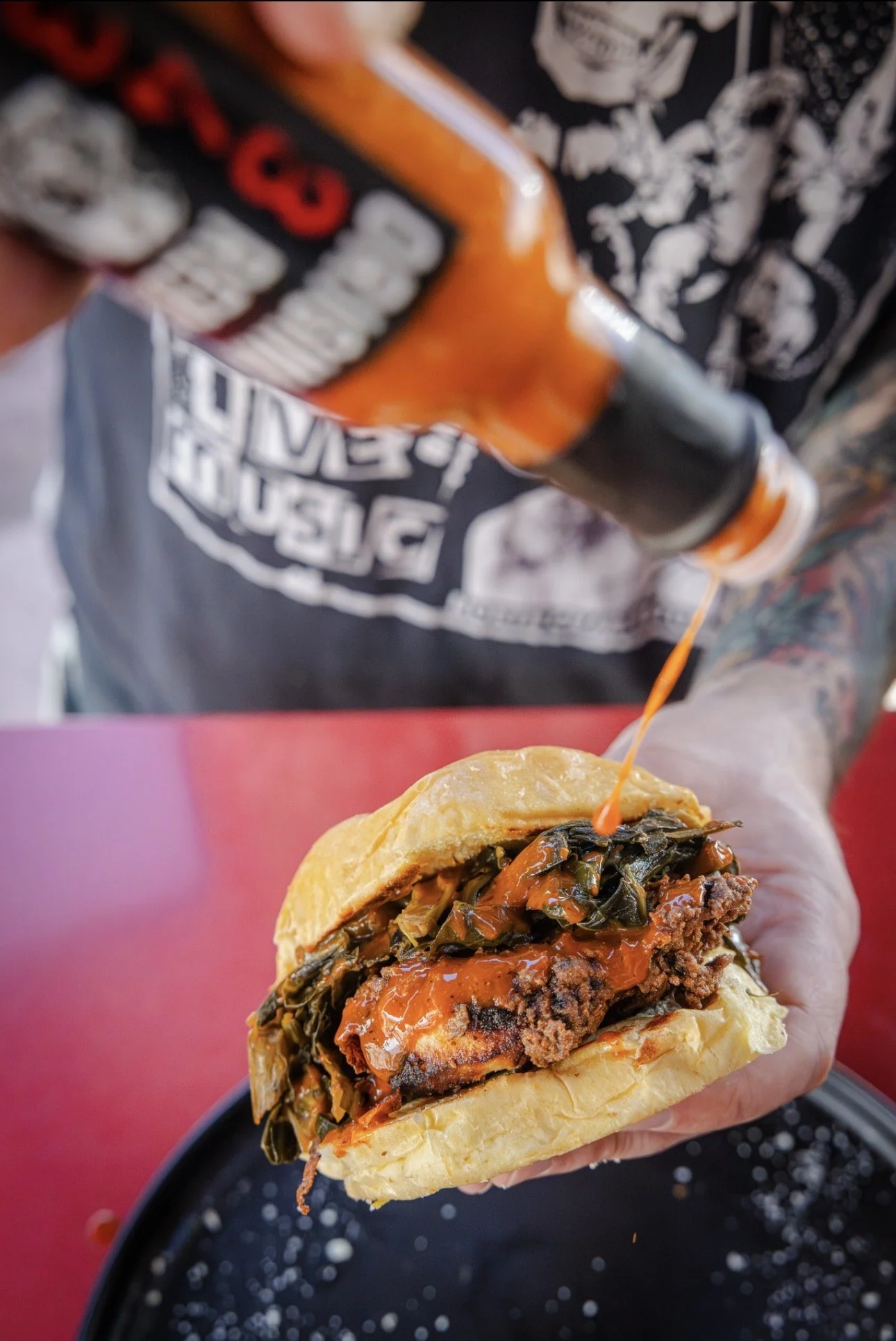 A person holding a sandwich filled with cooked greens, meat, and sauce, with a bottle of sauce being poured onto it.