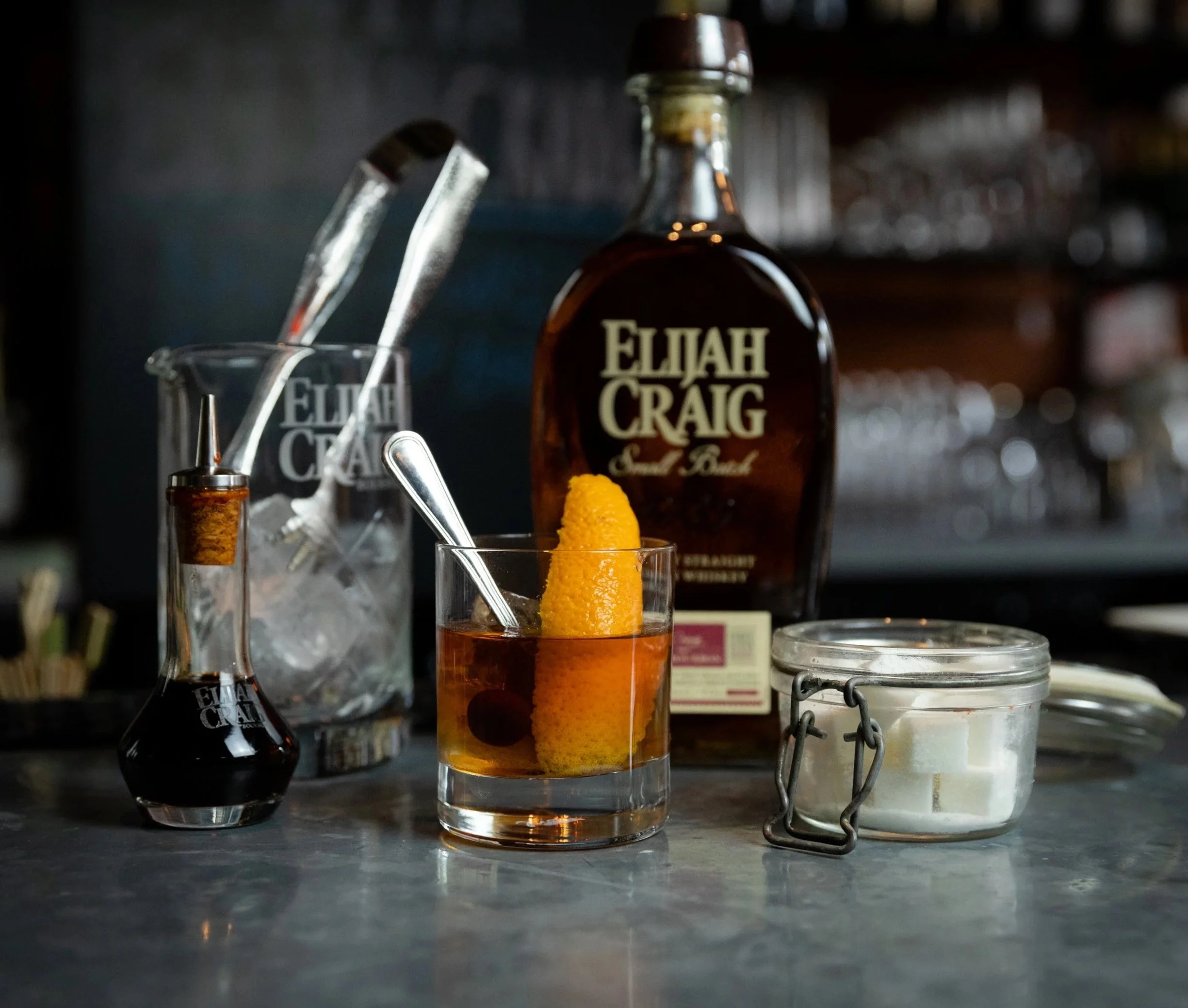 Close-up of Elijah Craig bourbon bottle, a glass with an orange peel garnish, and bar tools on a dark countertop in a bar setting.