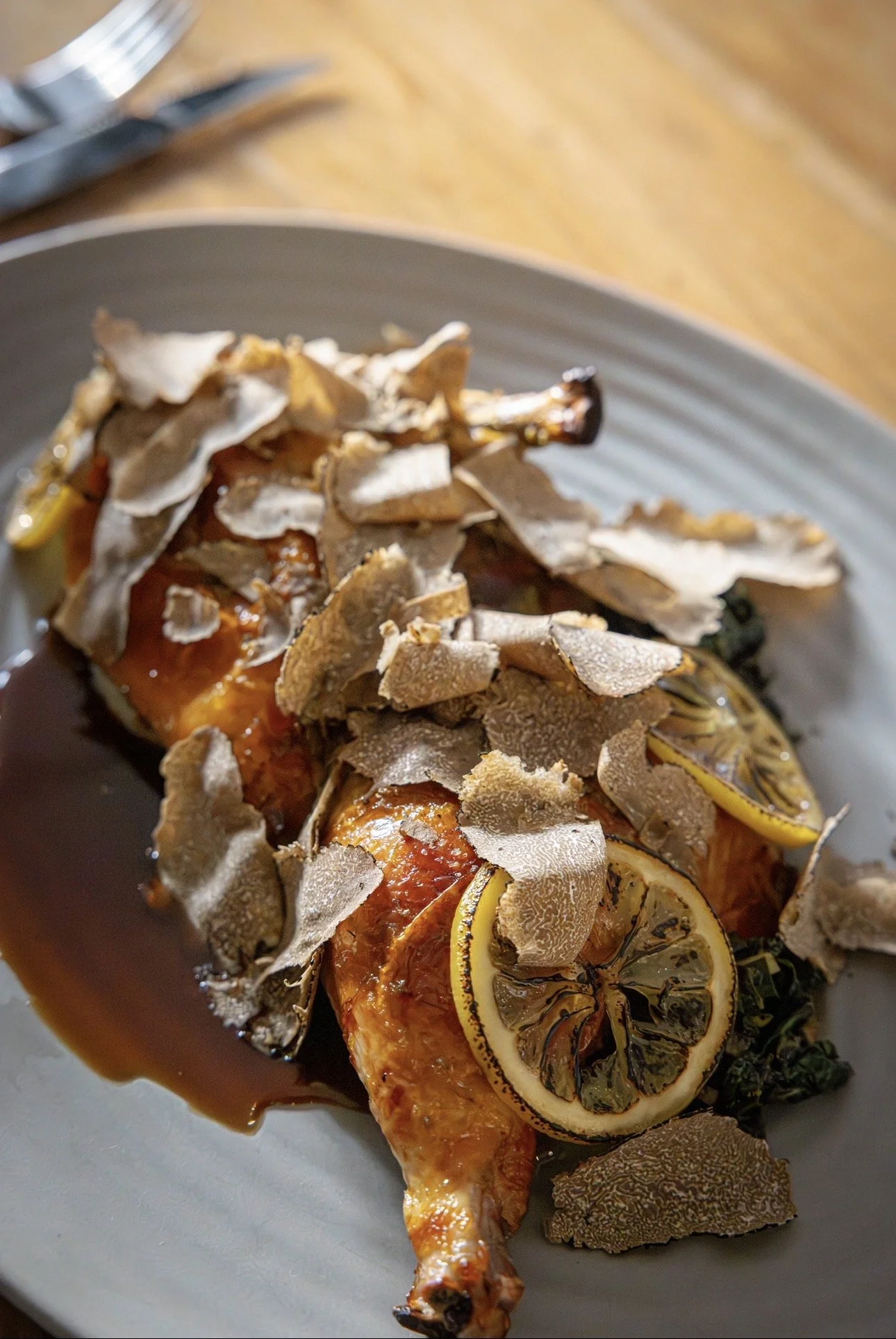 Roasted chicken leg garnished with lemon slices and topped with sliced truffles, served with sauce on a gray plate.