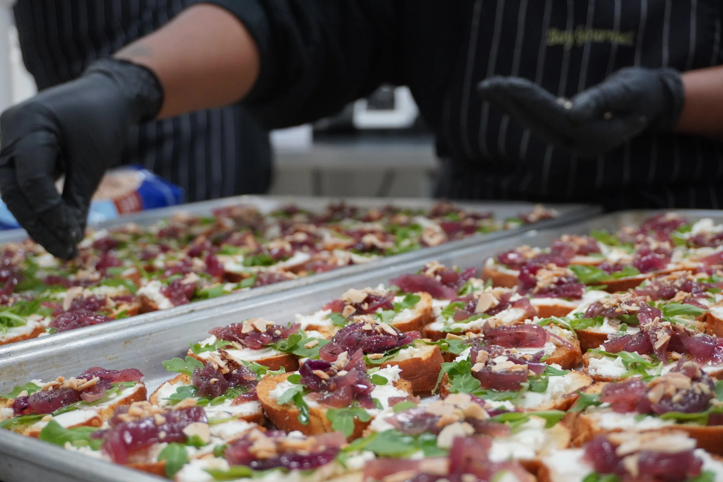 A person preparing slices of bread topped with ingredients including green arugula, sliced meat, cheese, and possibly nuts, arranged on a large tray.
